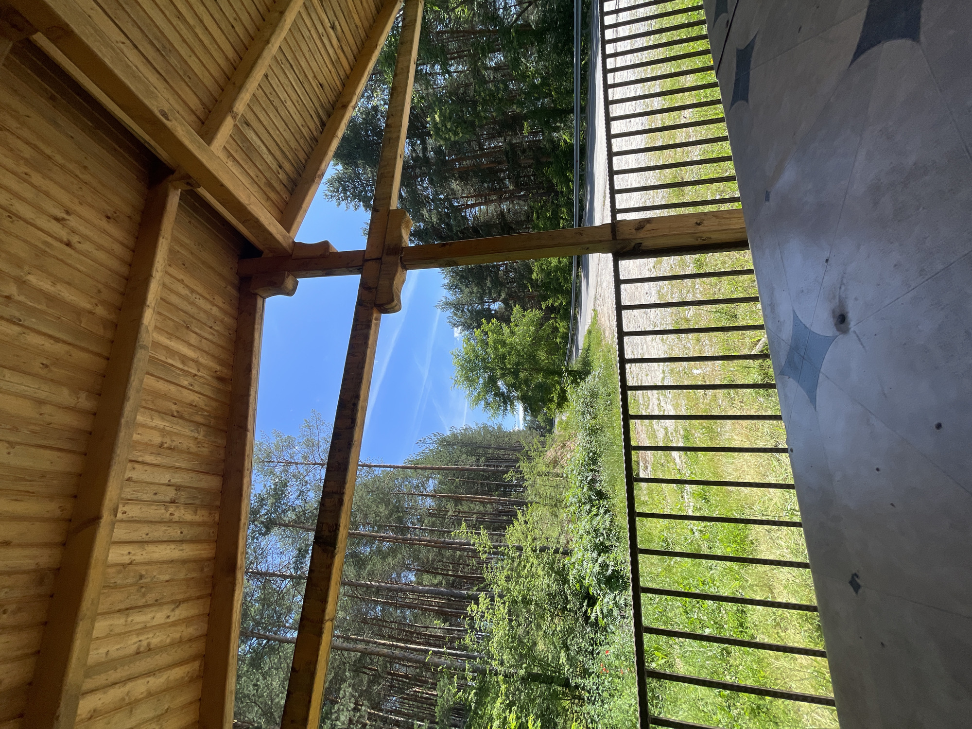 The image depicts an outdoor wooden structure with a slatted ceiling and exposed wooden beams, likely a pavilion or gazebo. Through an opening in the structure, a view of a forest with tall pine trees is visible under a bright blue sky with sparse white clouds. A paved road or pathway is seen in the middle distance, winding through the trees. In the foreground, a black metal railing separates the structure from a grassy area. The ground beneath the railing is covered in green grass and some low-lying plants. The front of the image shows a portion of a patterned surface, possibly a table, made of a light-colored material with dark gray geometric designs. The scene appears to be a tranquil natural setting, possibly a rest stop or recreational area, located in the vicinity of Valkosel, Bulgaria. No people are visible in the image.