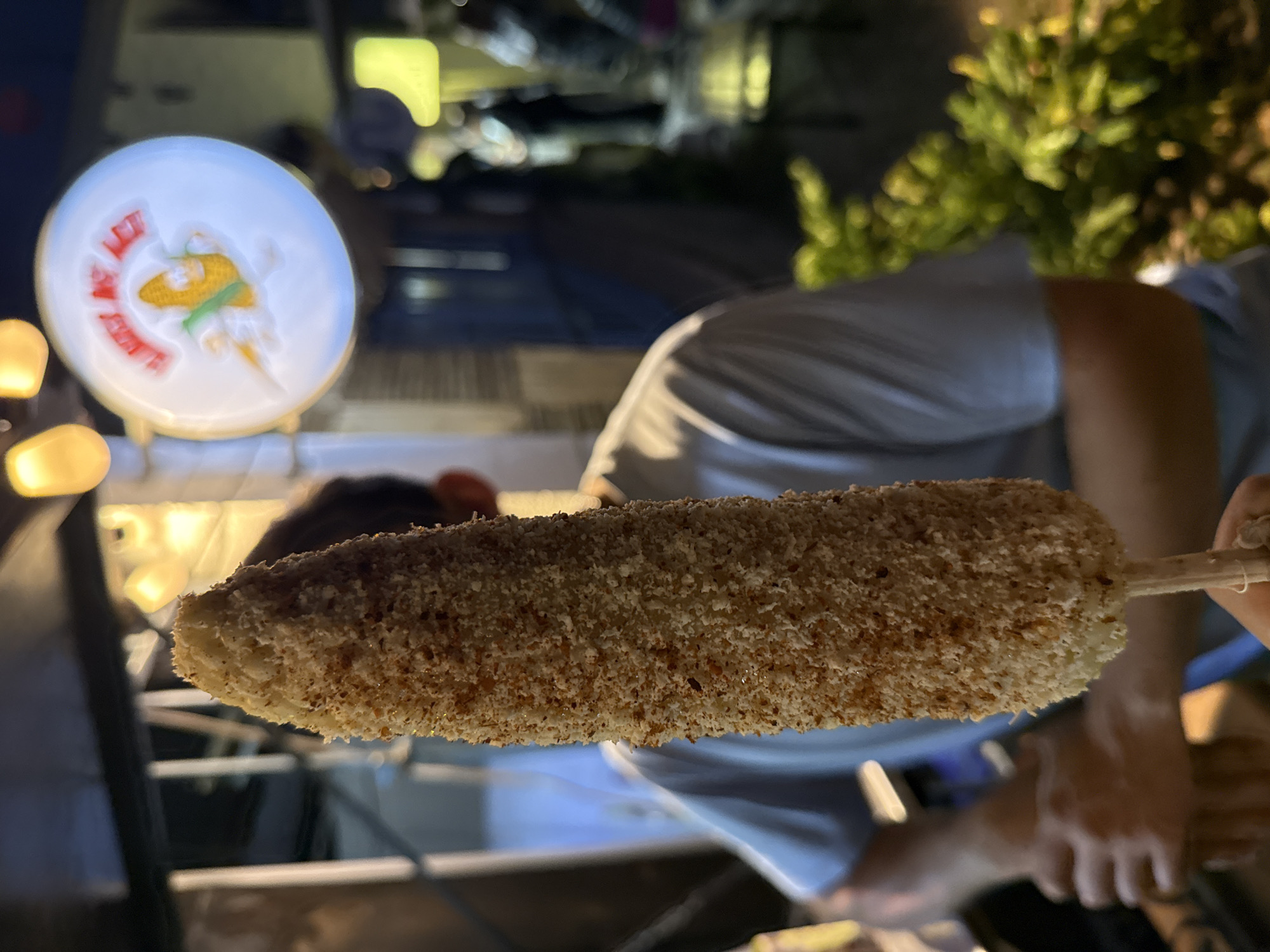 An eye-level, medium shot shows a hand holding elote on a stick, a common street food in Mexico, with the logo of the "Elotes Puebla" cart visible in the background. The elote is coated in a breading, possibly parmesan, and is the clear focal point. The hand holding the elote appears to be attached to a person wearing a light gray shirt, but their face is obscured behind the corn. The Elotes Puebla cart has a white sign with a stylized image of corn and the establishment's name written in red and white. The background is a blurred urban scene at night with artificial lighting and some greenery.