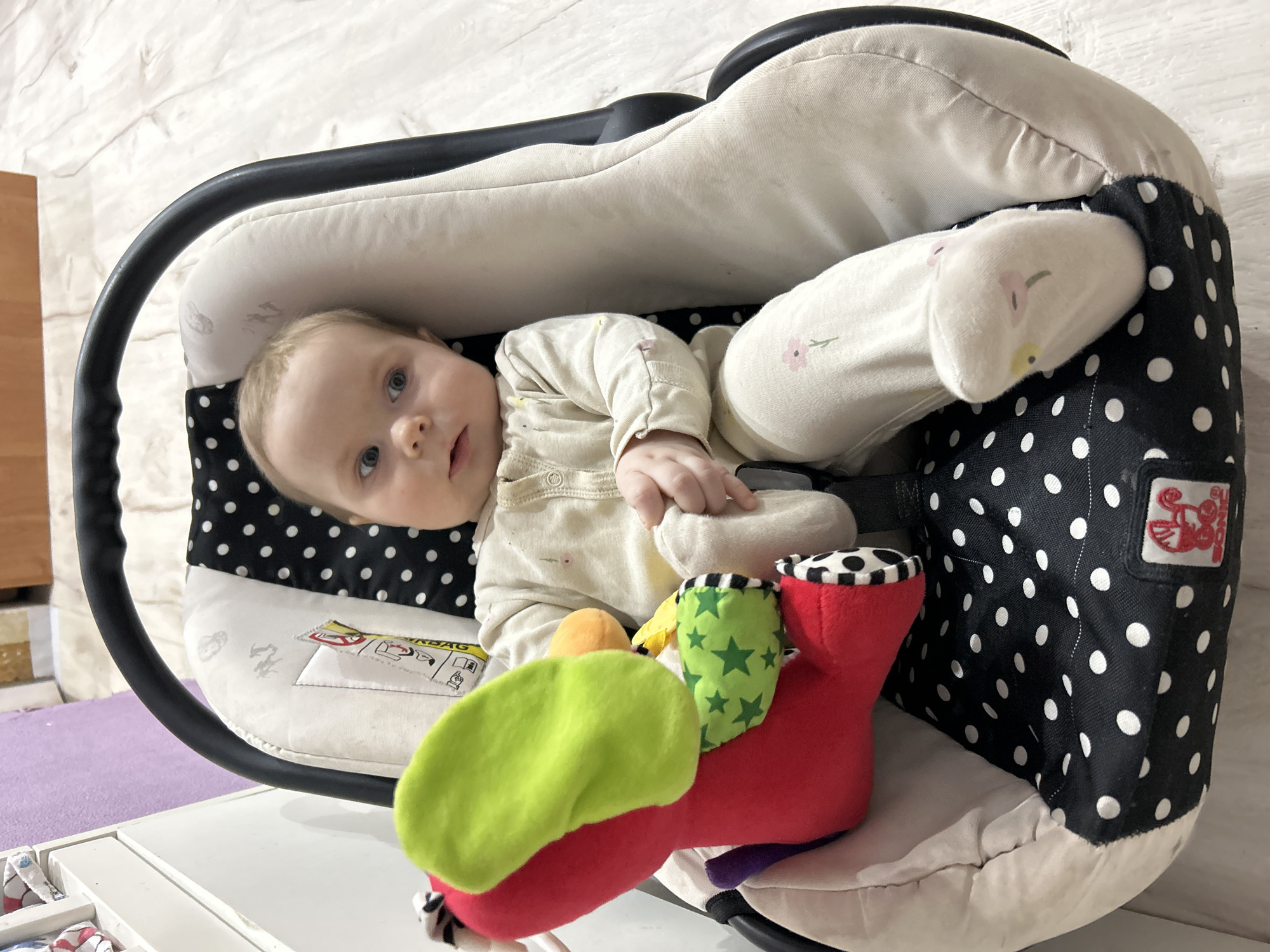 A baby is lying in a black and white polka-dot car seat, with a multi-colored plush toy at its feet. The baby is wearing a light beige, full-body outfit with small floral patterns on the legs. The surrounding environment appears to be indoors, with a textured white wall in the background. The location in the city of Rusalevka, Russia, is not identifiable from the image.