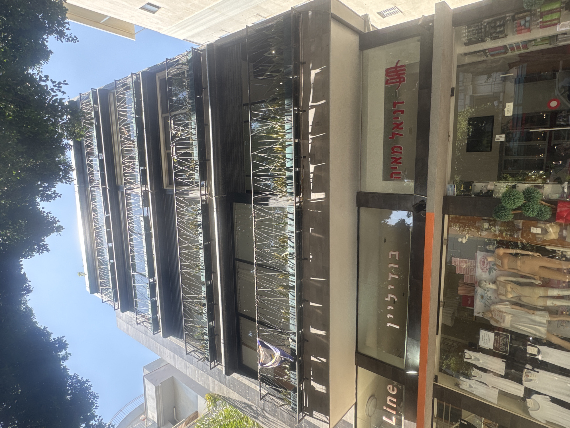 The image shows a building in Tel Aviv, Israel, viewed from a low angle. The building has a modern facade with multiple floors and balconies featuring decorative metal railings. The storefront displays mannequins dressed in white clothing, visible through the glass. The signs visible on the store are in Hebrew. Trees are visible on the sides of the image, suggesting an urban setting. The sky is blue.