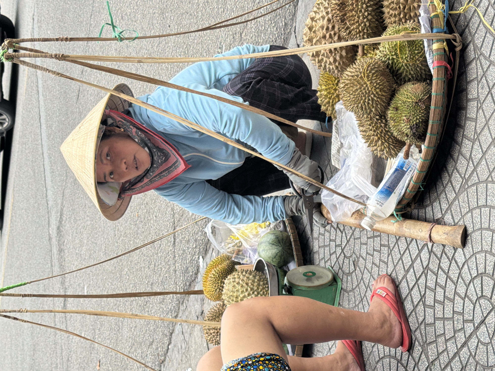 A woman wearing a conical hat and a blue jacket is squatting on a sidewalk in Thuận Hóa Ward, Vietnam, next to a collection of durian fruit and a green scale. She is wearing gloves and appears to be tending to the fruit. To her left, the legs of another person are visible, wearing colorful shorts and red sandals. The durian fruits are arranged in a basket, with some on a scale and others on the ground. The sidewalk is paved with a circular pattern of gray tiles. In the background, the street is visible, with a car and trees. The overall scene depicts a street vendor preparing to sell durian fruit.