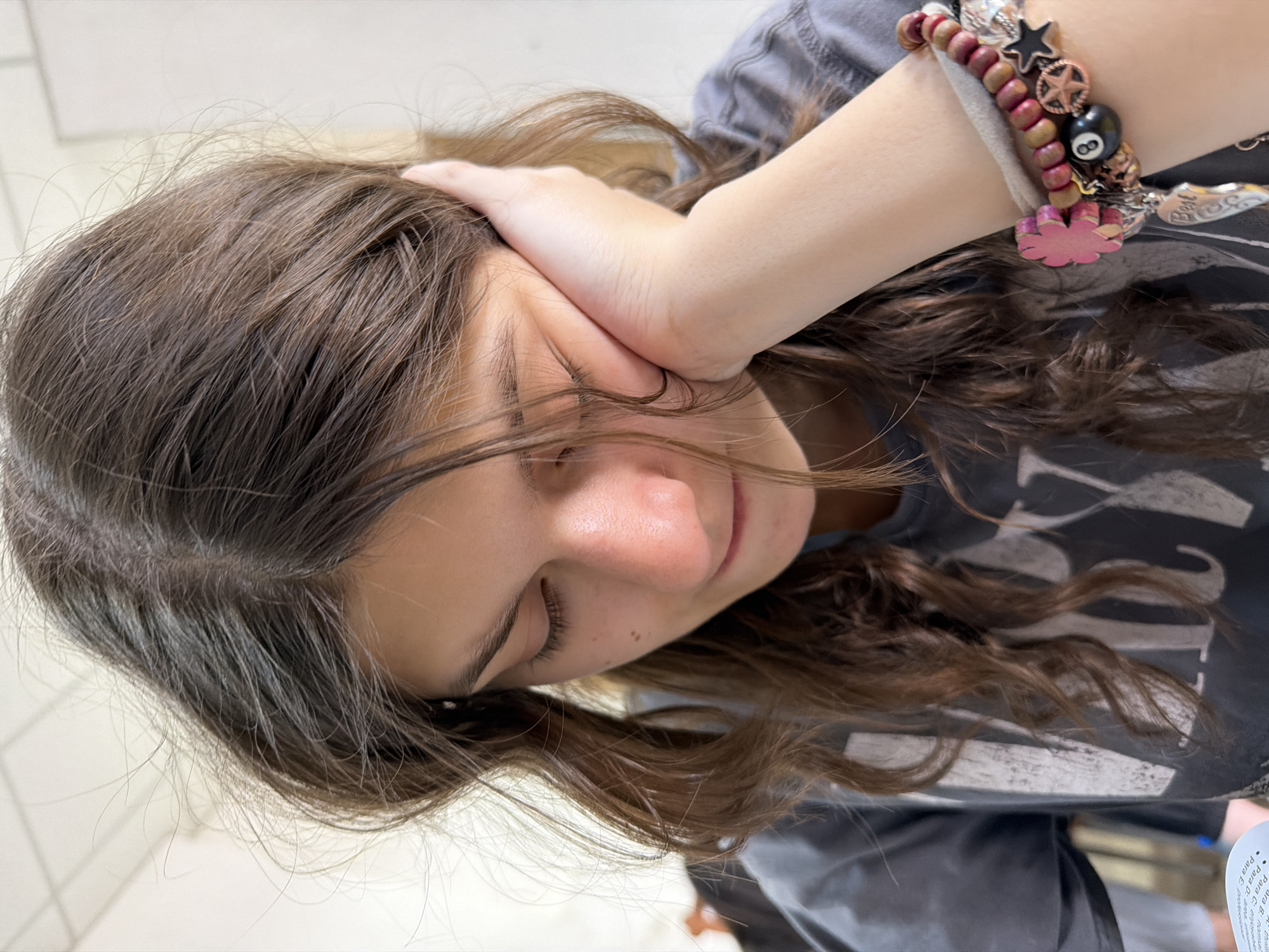 A close-up shows a young woman with brown hair, her head tilted, resting her hand on her face. She is wearing a grey t-shirt and a beaded bracelet with charms. The background is out of focus, revealing only light, indistinct surfaces. There is no indication of the location or surroundings.