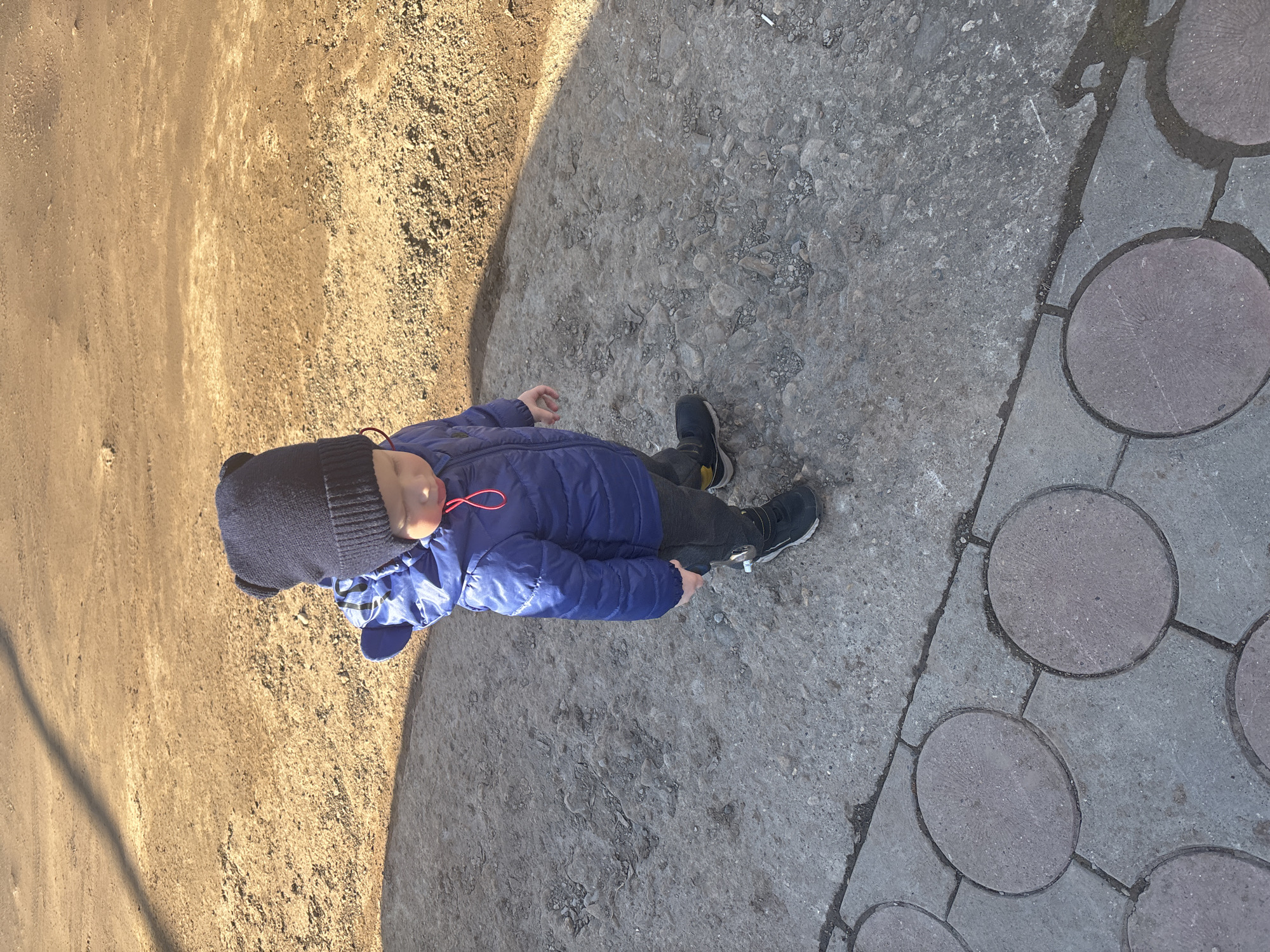 A child wearing a blue winter coat and a dark beanie stands on a textured concrete surface in what appears to be an outdoor area. The surface transitions to a dirt area on the left and a patterned walkway made of round pavers on the right. The location is the city of Ферма, Russia. The child is looking down.