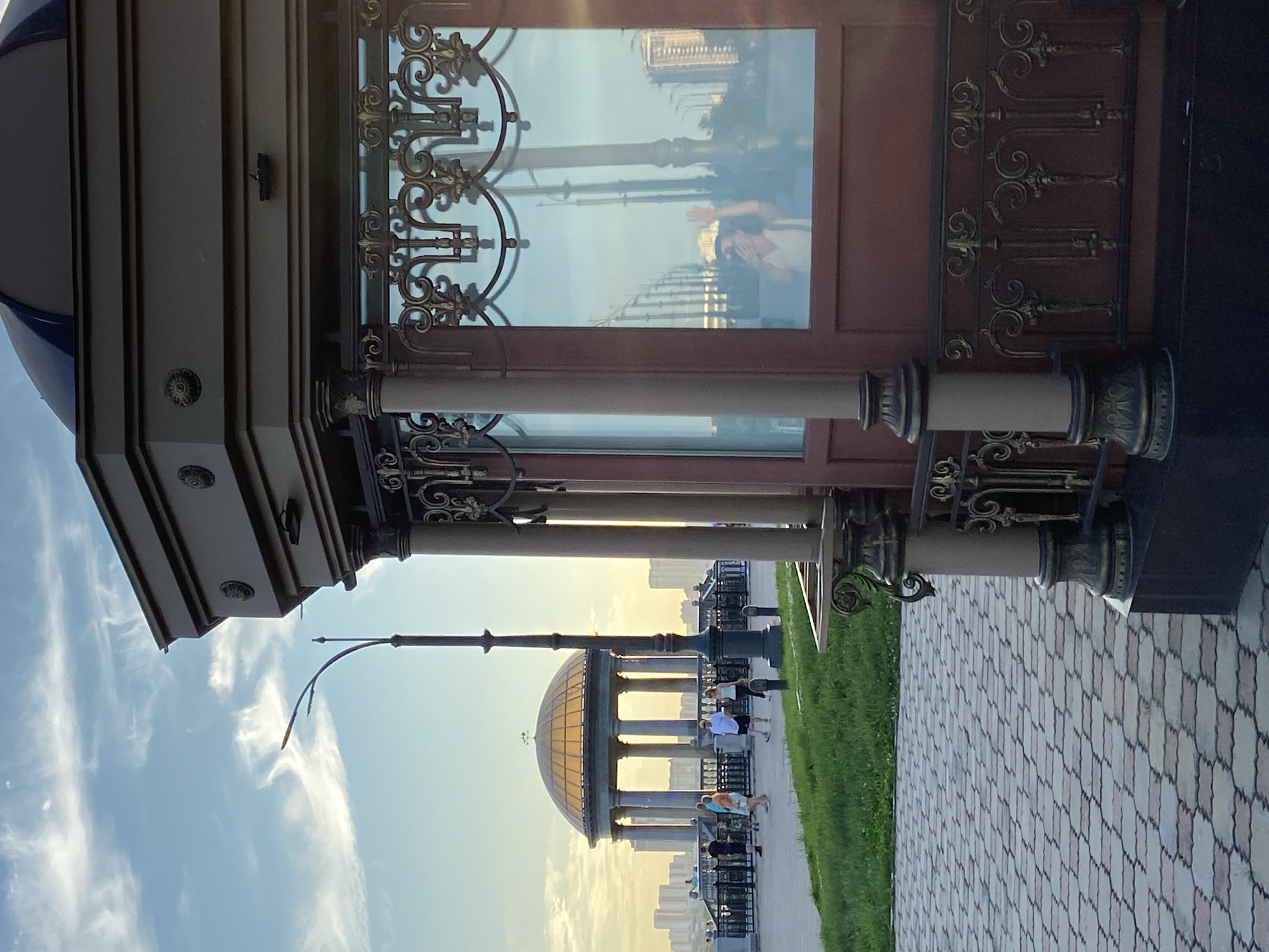 This is a medium shot, eye-level, outdoor photograph taken in Blagoveshchensk, Russia. The image captures a view of a promenade along the Amur River at sunset. In the foreground, on the right, is the corner of a dark red, ornate pavilion with decorative metalwork and columns. Its glass windows reflect the scene outside. A woman is visible in the reflection, taking a selfie with her phone and waving. To the left of the pavilion, a paved walkway with a geometric pattern of light and dark stones leads towards a grassy embankment. A lamppost with a curved arm stands between the pavilion and the walkway. In the background, across the Amur River, the city skyline of Heihui, China, is visible with numerous buildings. A classical-style rotunda with a domed roof and columns is situated on the promenade, with a railing in front of it and several people walking along the path. The sky above is a clear blue with wispy white clouds, and the setting sun casts a warm glow on the scene. The environment appears to be a public park or waterfront area.