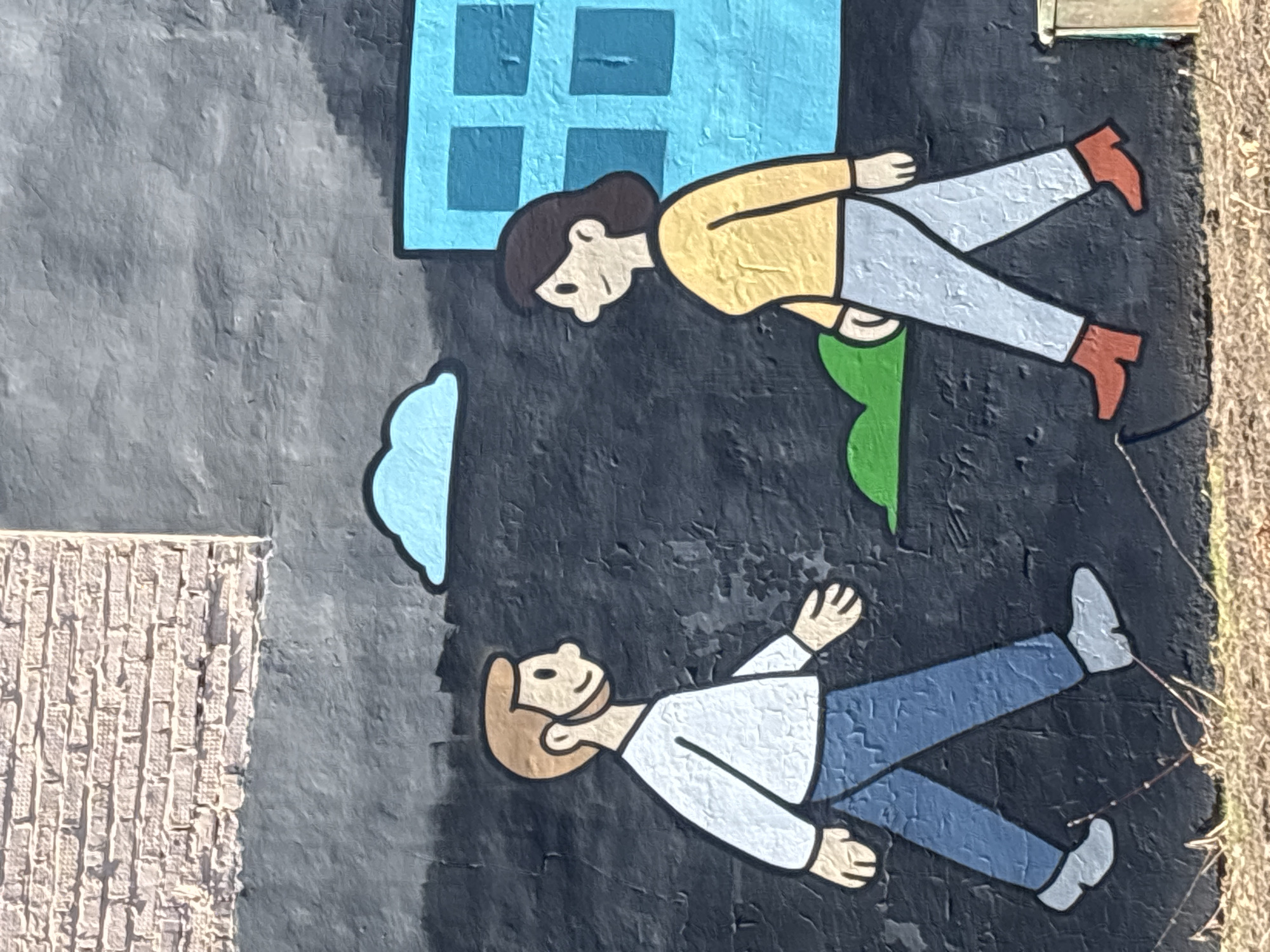 The image depicts a mural in Izhevsk, Russia, featuring two cartoon-like figures against a textured, dark background. The upper figure, positioned horizontally, has brown hair, a yellow shirt, and light gray pants with red shoes. A green shape is located below them, and a blue window is above.
The lower figure, also horizontal, has tan hair, a white shirt, blue pants, and light gray shoes. To the left of the figures is a gray brick-like texture, resembling a wall or building. To the right of the lower figure is some dry grass.