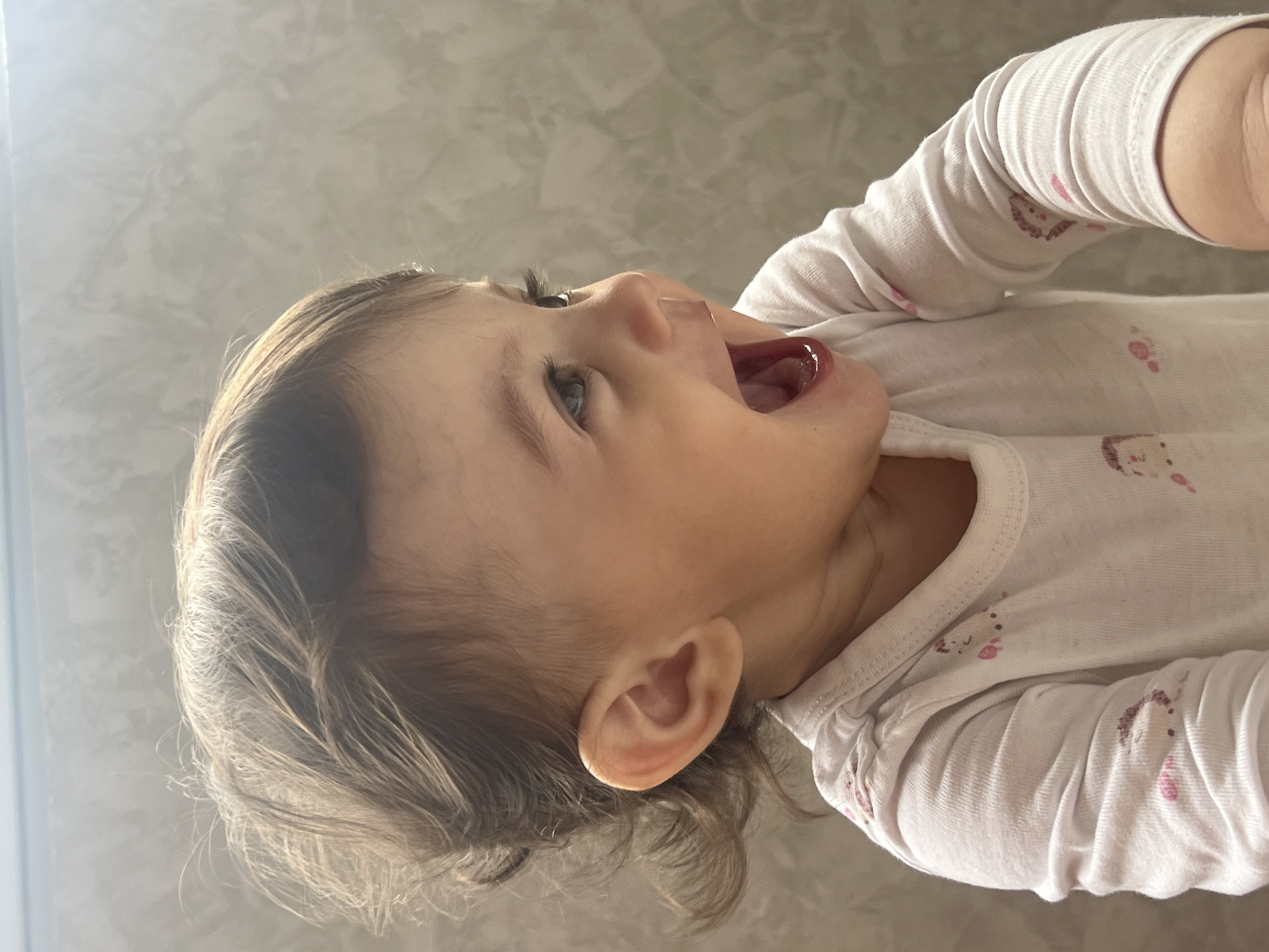 A close-up, rotated, eye-level shot depicts a baby girl with light skin, brown hair, and blue eyes looking upwards with her mouth open. The baby is wearing a white long-sleeved shirt with a pink unicorn pattern. The background consists of a blurred, light gray surface. There is no evidence of Elena, Bulgaria.