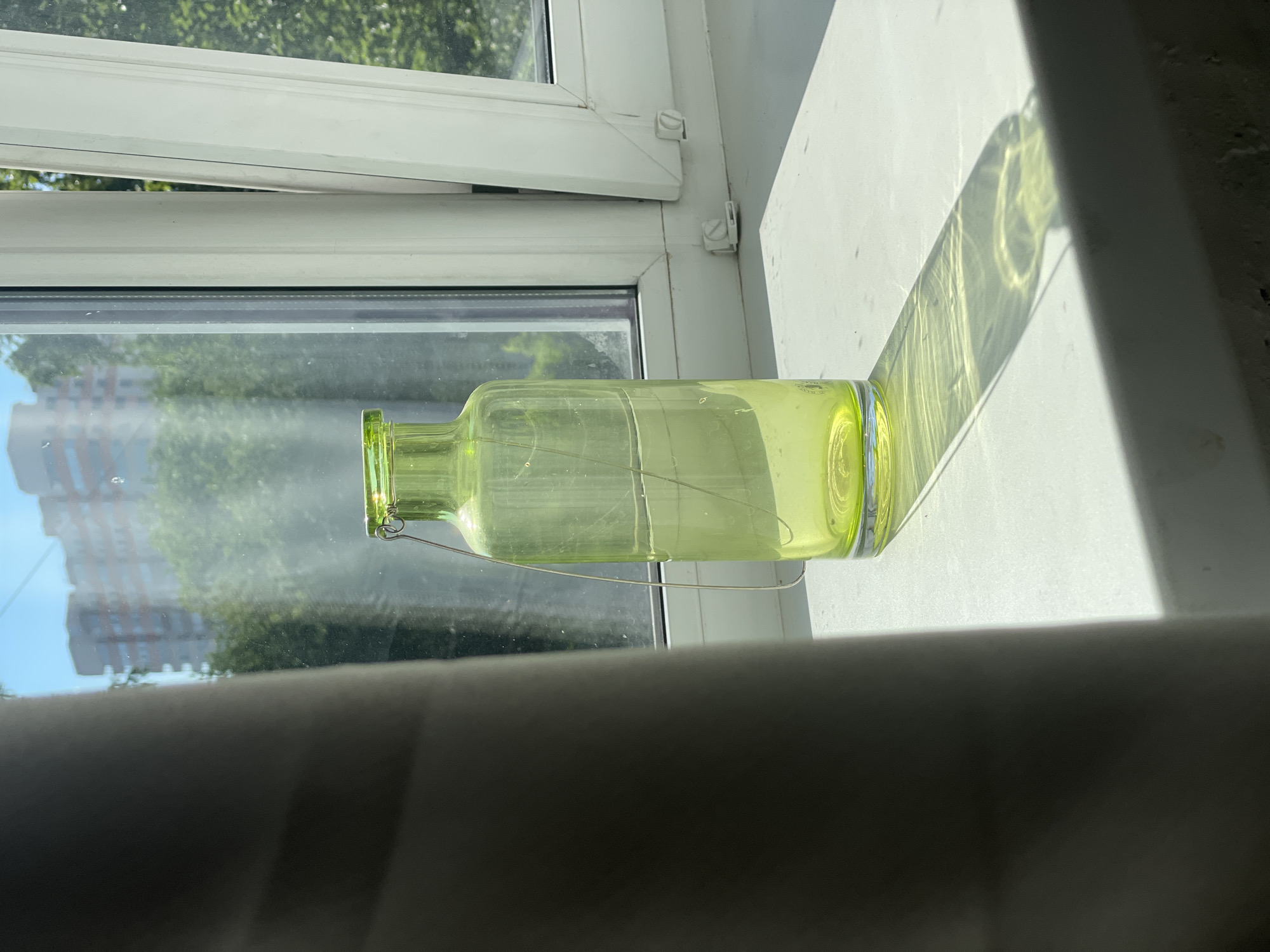 A light green glass bottle with a thin wire handle is placed on a white windowsill in front of a window. The bottle is partially obscured on the left by a dark grey curtain. Through the window, trees and a multi-story building are visible against a bright blue sky. Sunlight casts a long shadow of the bottle onto the windowsill. The scene is set indoors, on what appears to be a sunny day. There are no people or animals present. The environment suggests a residential setting.