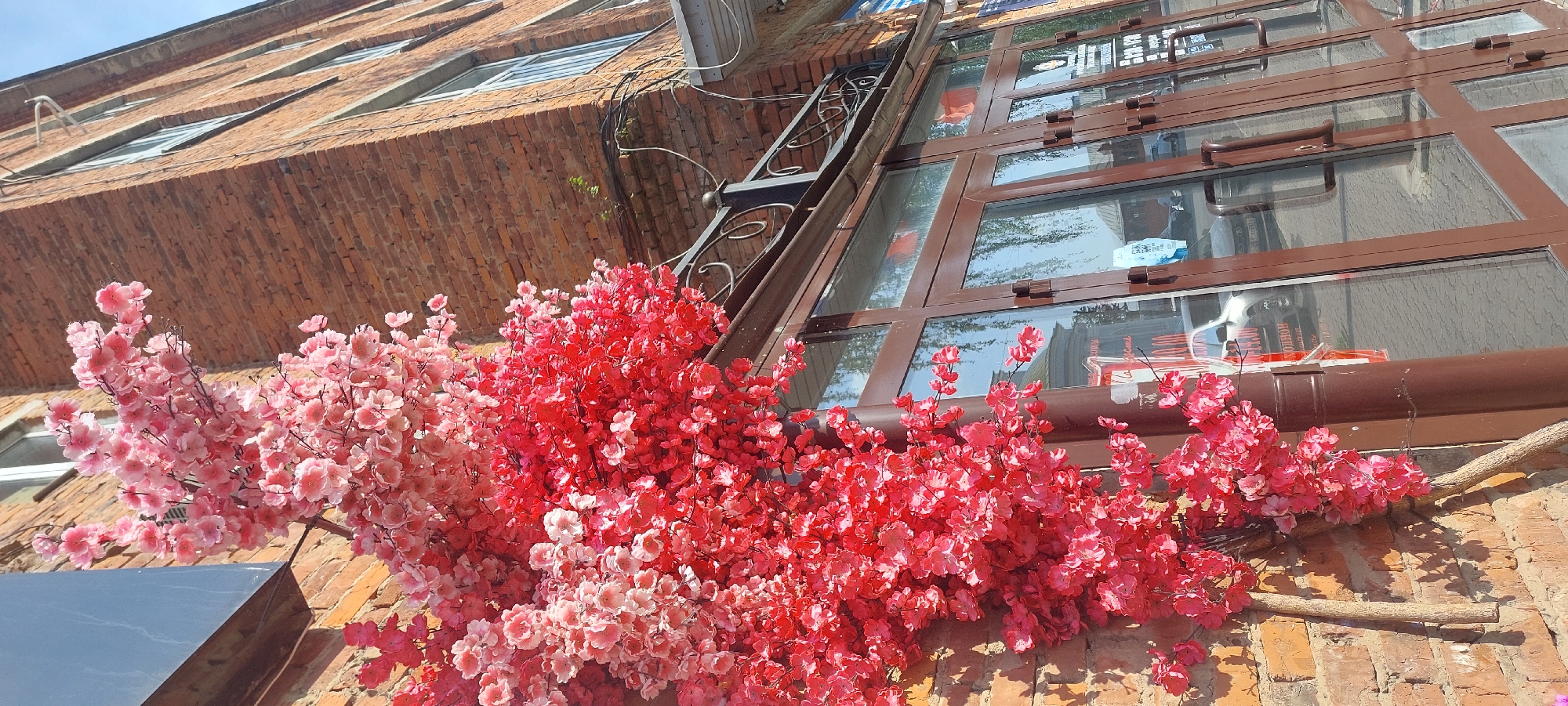 A low-angle, full-shot photograph depicts a facade of a building with red brick walls and large, brown-framed glass doors. The building appears to be in Votkinsk, Russia, given the presence of Russian text on posters visible on the glass doors. Attached to the brick wall on the left is an artificial tree with numerous bright pink and red blossoms, creating a vibrant display. Two wooden branches extend from the base of the artificial tree, leaning against the wall. The glass doors reflect the sky, overhead wires, and some buildings across the street. A black metal railing with decorative scrolls is positioned above the glass doors, attached to the brick wall. The sky is clear and blue.