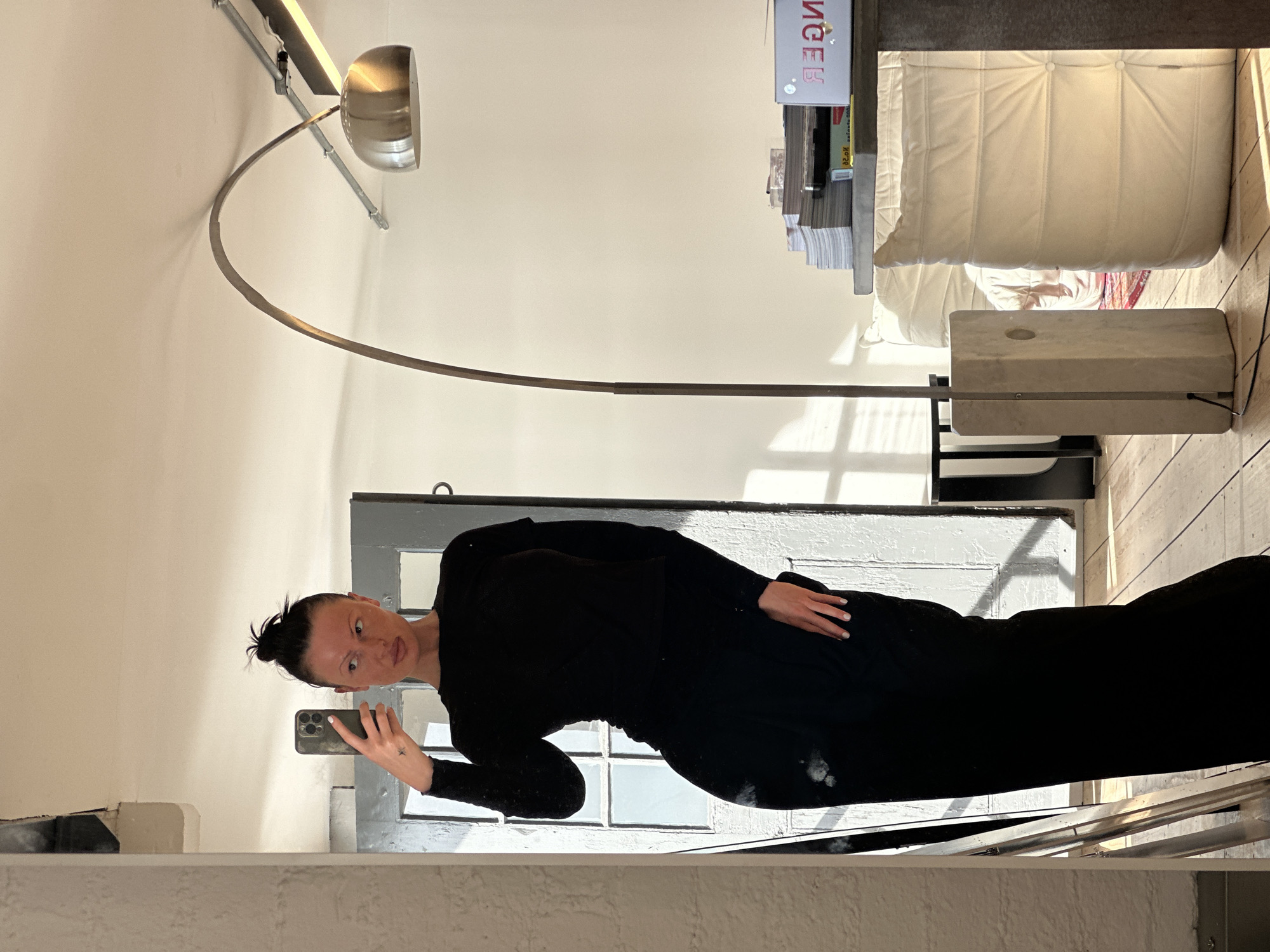 An adult female, reflected in a large mirror, is taking a selfie with a dark-colored smartphone. Her dark hair is pulled into a bun, and she wears a long-sleeved black top and black trousers. A small star tattoo is visible on her left forearm. The interior setting features light wooden floorboards and a white-painted brick wall on the left. Behind the mirror, a light grey, multi-paneled door with glass inserts is visible, receiving direct sunlight that casts distinct shadows. A large, arching floor lamp with a metallic dome shade stands on the right, originating from a light-colored rectangular base. To the right, a large, quilted cream-colored upholstered object, possibly a sofa section or ottoman, is present. A dark table or counter behind the lamp holds stacks of books and magazines. The architectural style suggests an urban residence or studio within London, United Kingdom, consistent with renovated industrial or commercial properties commonly found in areas such as Shoreditch or Clerkenwell.