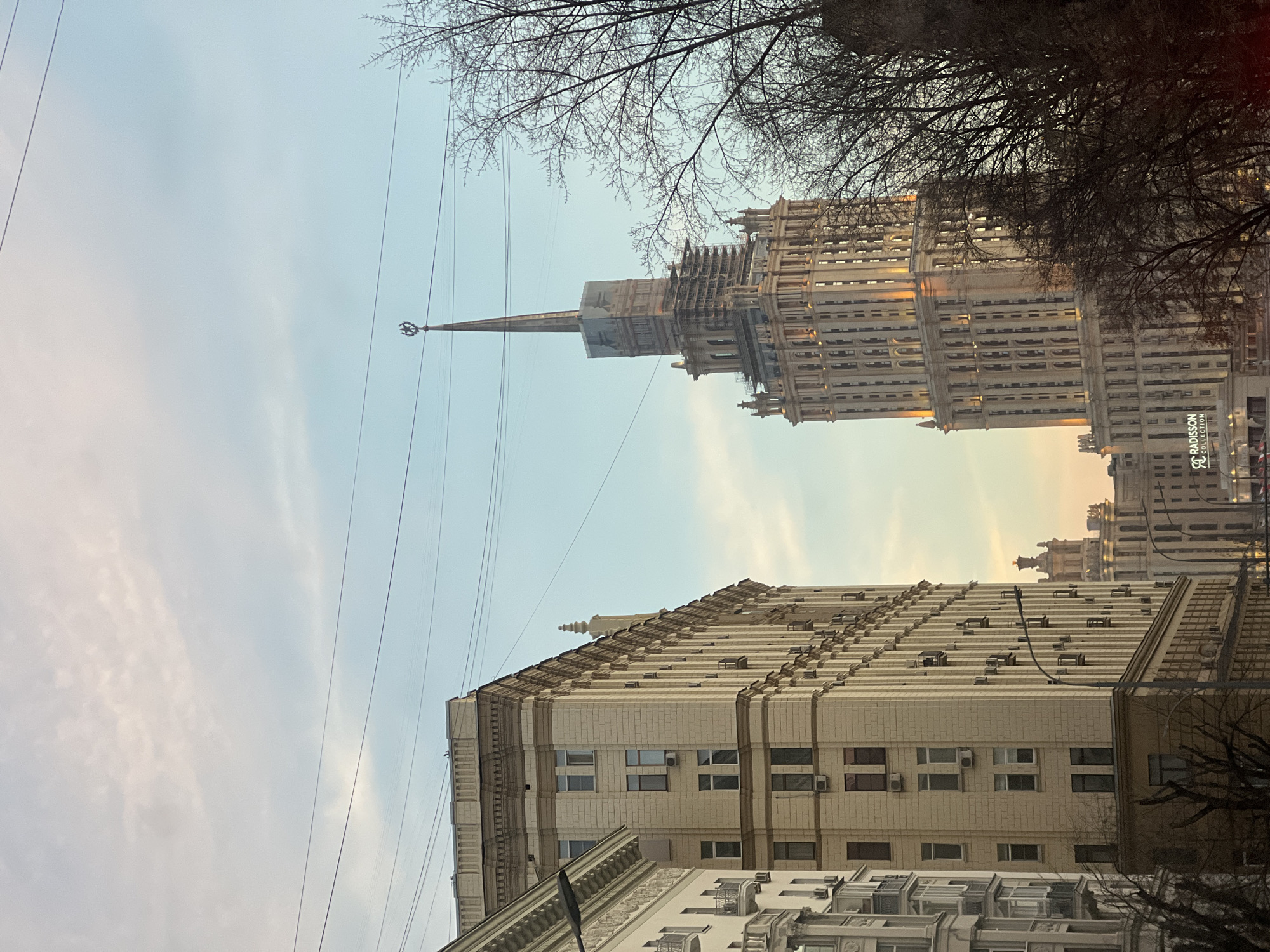 The image displays a Moscow street scene featuring the Radisson Collection Hotel (formerly Hotel Ukraina) on Kutuzovsky Avenue. The hotel, a prominent Stalinist-era skyscraper, occupies the right side of the frame. It presents a light-colored, multi-story, ornate facade, with scaffolding evident on its upper sections, culminating in a star-topped spire. The "Radisson Collection" branding is visible on its lower portion. To the left, a multi-story building of similar architectural style features a beige facade with numerous windows, many equipped with external air conditioning units. Bare deciduous tree branches are visible in the lower right and left foregrounds. Multiple dark utility wires crisscross the sky, which exhibits a gradient from light blue to a pale orange-pink. No discernible human figures or vehicles are present.