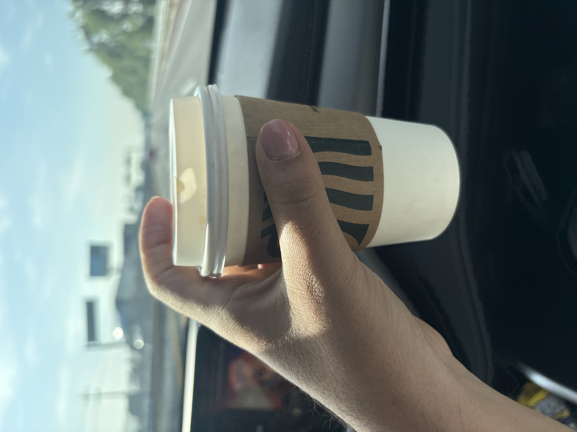 A hand is holding a Starbucks coffee cup with a sleeve in a car. The scene is captured from inside a vehicle, looking outward toward the exterior environment. The background shows a blurred view of what appears to be a street or highway, with trees, buildings, and vehicles visible under a bright sky. The photo was most likely taken in a car and not from San Lucas el Grande, Mexico. The hand holding the coffee cup is light-skinned, and the fingernails appear to have pink nail polish. The coffee cup is white with a brown Starbucks sleeve, and a white lid. The interior of the car is visible, with dark panels and a glimpse of the dashboard.
