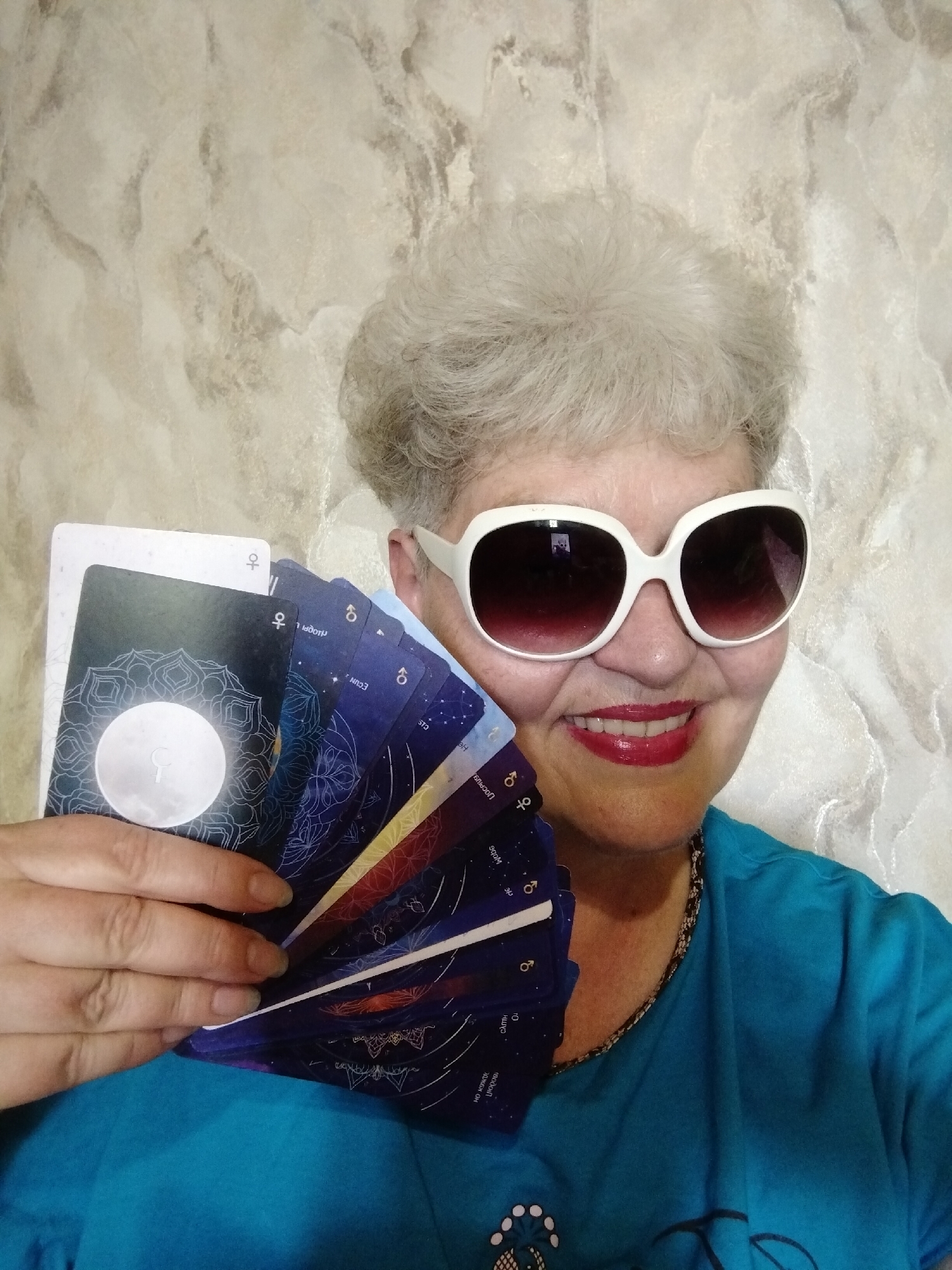 An older woman with short, wavy grey hair is featured in a close-up indoor portrait. She wears large white-framed sunglasses with dark lenses and bright red lipstick, displaying a cheerful, confident smile. She is dressed in a teal t-shirt with a patterned neckline. Her left hand holds a fan of dark-colored oracle or tarot cards, prominently displaying celestial and mandala-like designs. One card features a large white moon with a crescent symbol. The background is a light-colored, textured wall with a marble-like pattern. The location is Horishni Plavni, Ukraine.
Visible text on the cards, as identified by OCR, includes: "우" (likely a symbol for Venus/female), "8" (likely a symbol for Earth or an astronomical node), "дот", "ECUN", "CIS", "JOCPIUS", "He", "900M", "光", "10", "HEYD", "водой", and "ОДЖБХ ОН".