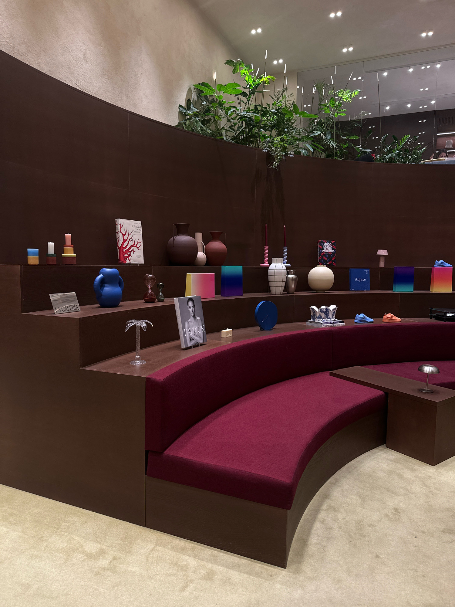 The image depicts the interior of a retail store in Madrid, Spain. The store features a tiered display area with dark brown wooden shelves showcasing various decorative objects, books, and plants. The shelves are arranged in a semi-circular fashion, with a curved, deep red upholstered seating area in front of them. The walls are a textured off-white color.
On the shelves, notable items include stacks of colorful candles, several vases of different shapes and sizes, books with prominent covers, and decorative objects such as a blue ceramic vase, glass hourglasses, and a clear palm tree sculpture. On the lower shelves, two pairs of athletic shoes, one blue and one orange, are displayed. A small, silver lamp with a dome-shaped shade sits on a small, dark brown side table next to the seating area. In the background, there are glimpses of lush green plants, reflective surfaces, and ceiling lights. No people are clearly visible in the frame. The overall impression is one of a modern, artfully curated retail space.