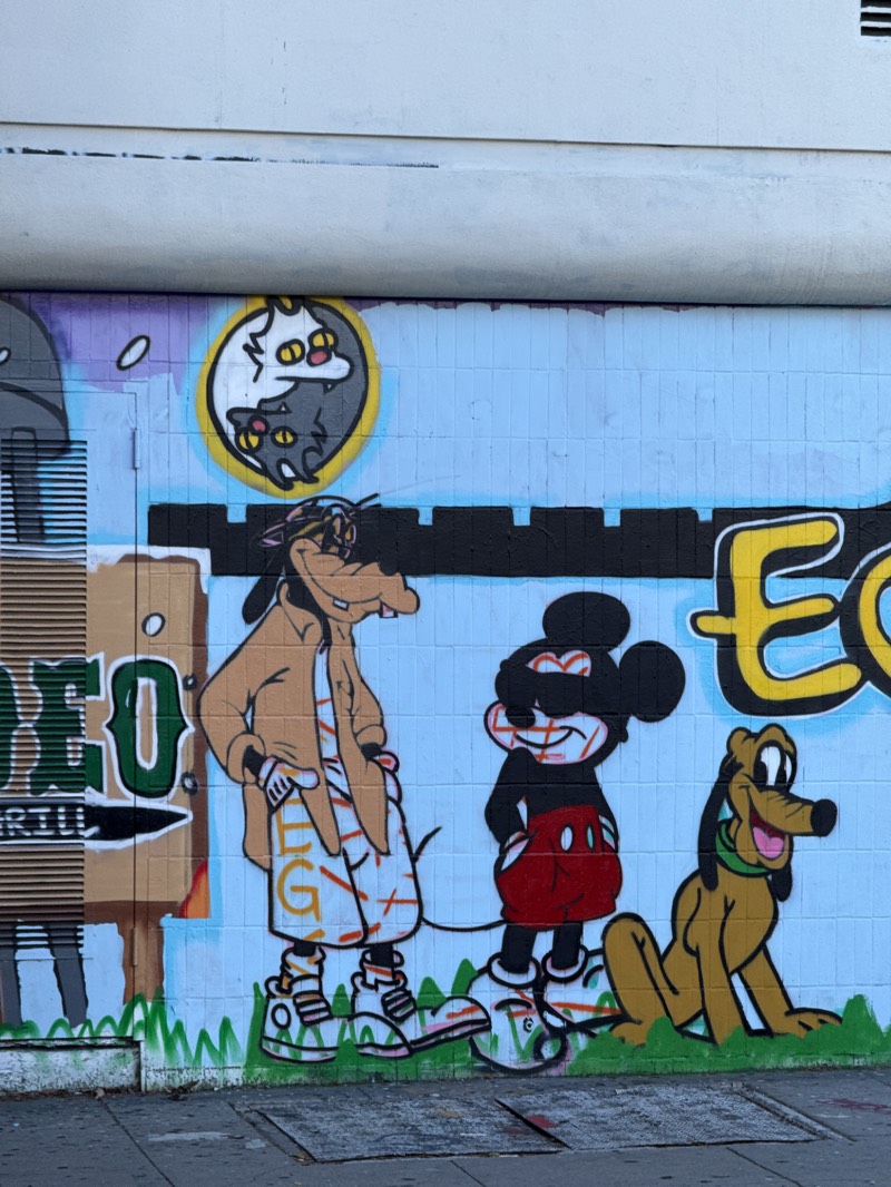 A graffiti mural on an exterior wall in an urban environment depicts cartoon characters including Goofy, Mickey Mouse and Pluto, standing on a patch of painted green grass. Goofy, depicted in tan jacket and shorts, stands to the left. Mickey Mouse is to the right of Goofy, wearing sunglasses and red shorts. Pluto is to the right of Mickey Mouse, seated. An image of yin-yang cats is above and to the left. Portions of other graffiti surround the characters. The scene is likely Los Angeles based on the graffiti and mural style.
