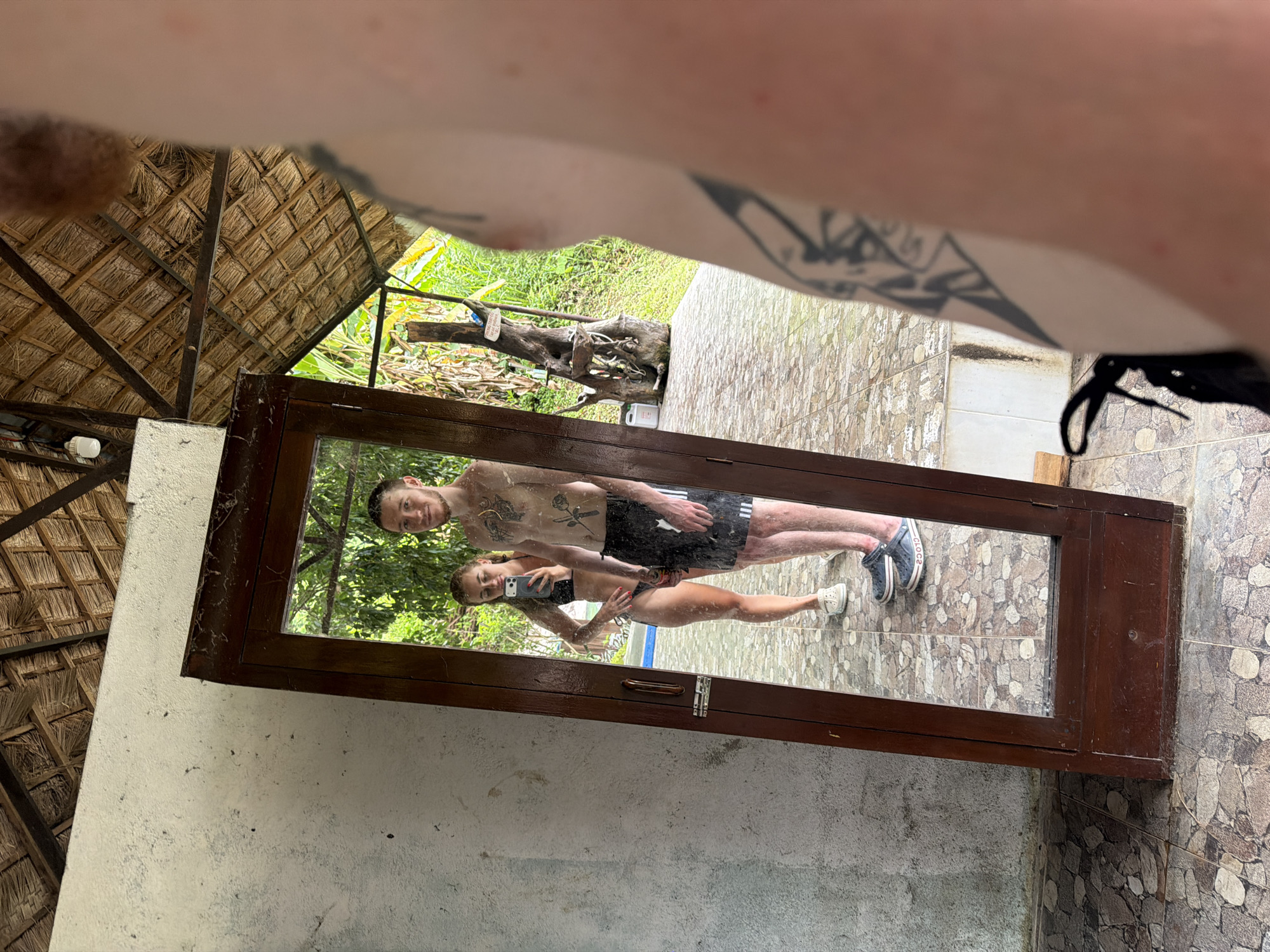 The media file shows a mirror reflecting a young couple. The woman is wearing a black bikini and the man is wearing black shorts. They are both shirtless and have tattoos. They are posing for a selfie in a tropical outdoor setting. The background of the reflection shows lush greenery, suggesting a warm climate. The mirror itself is framed in dark wood and appears to be part of a rustic structure, possibly an open-air changing room or bathroom. The setting has a thatched roof visible in the upper left portion of the frame. The overall mood is casual and relaxed. The floor in the reflection is tiled with a pattern of neutral-colored stones. The original image also captures parts of the environment outside the mirror, including the textured wall of the structure and the thatched roof, as well as a portion of the person holding the camera, whose arm with tattoos is visible in the upper part of the frame. The lighting suggests it is daytime, possibly morning or afternoon.