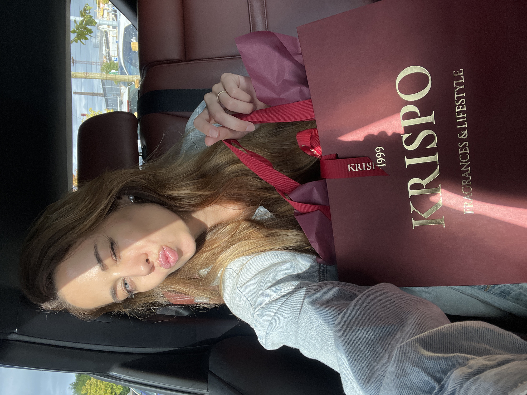 A young woman is sitting in the passenger seat of a car, taking a selfie. She is winking and puckering her lips. She is wearing a light blue denim jacket and a silver earring. In her right hand, she is holding a burgundy gift bag with red ribbons. The bag has "KRISPO 1999 FRAGRANCES & LIFESTYLE" printed on it in gold letters. The interior of the car appears to be leather. Outside the car, there are trees and buildings visible, suggesting an urban environment. The lighting suggests it is daytime, with sunlight casting shadows on the woman and the gift bag. The scene is likely in Klaipėda, Lithuania, given the context of previous interactions.