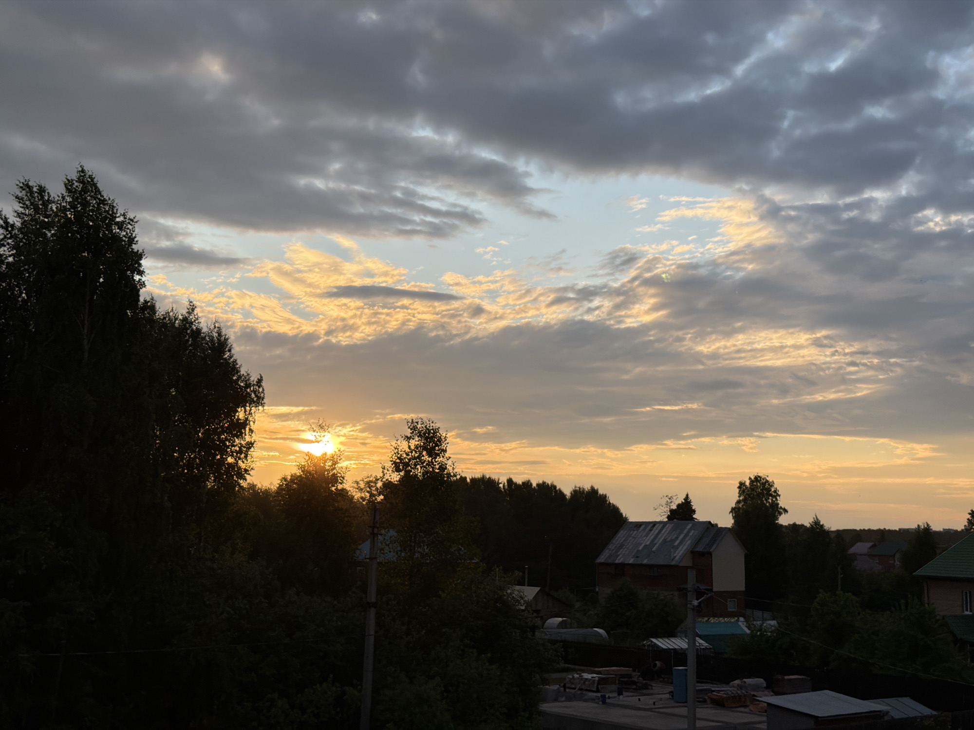 The image depicts a rural residential area at sunset, likely in Chaikovsky, Russia. The sky is filled with dramatic clouds, with patches of golden light breaking through, indicating the sun is low on the horizon. Silhouetted against the warm glow are numerous trees, some tall and dense, others smaller and more scattered.
In the middle ground, several houses are visible. One house, prominent on the right side of the image, has a red brick lower level and a lighter upper story, topped with a metal roof. Other structures, including what appear to be greenhouses and smaller outbuildings, are also present among the trees.
The foreground shows elements of what seems to be ongoing construction or storage, with various building materials and structures visible. A utility pole stands vertically on the left side of the image. The overall impression is one of a quiet, lived-in community on the outskirts of a town, bathed in the soft, fading light of the day. There are no people visible in the scene.