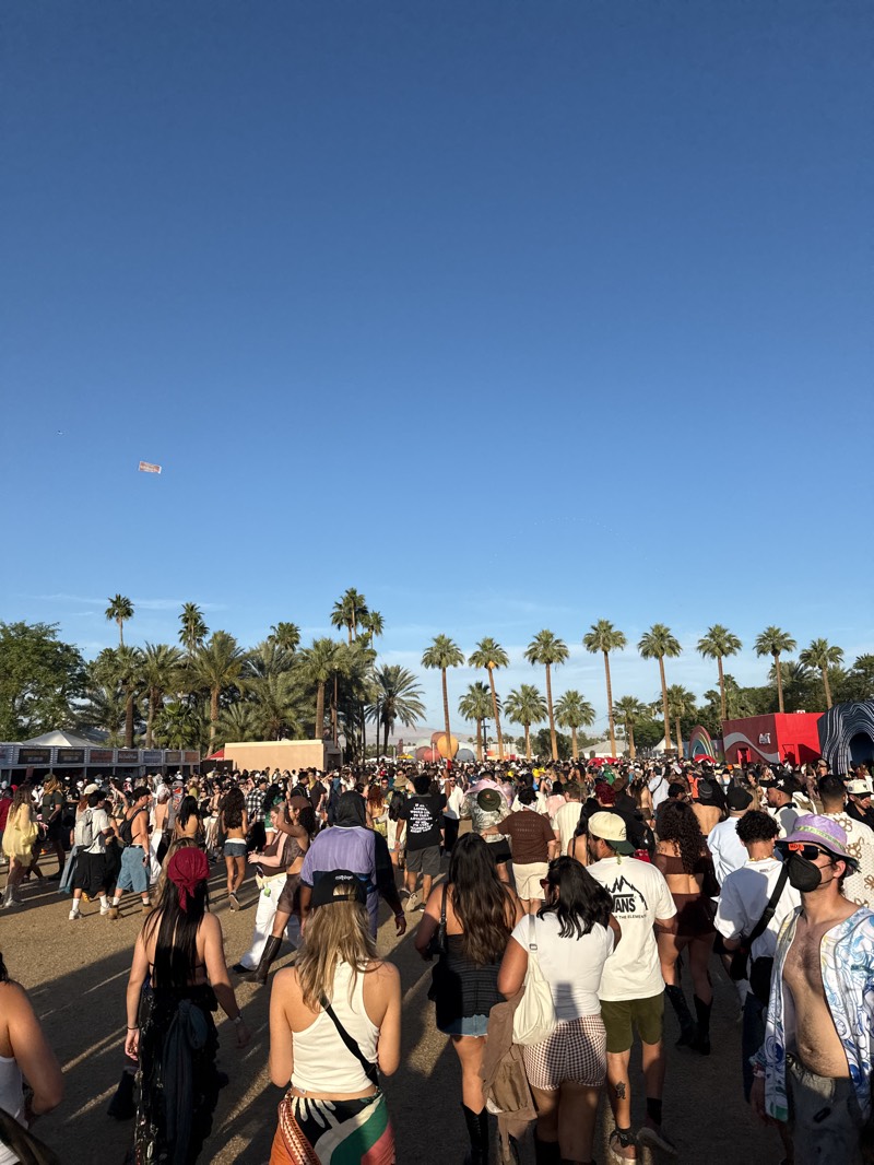 A large crowd of people is milling around in an open area in Indio, California, with palm trees lining the background. The sky is clear and blue, with a banner flying in the distance. The individuals in the crowd are wearing a variety of casual clothing, including shorts, tank tops, and hats. There are tents and structures visible in the distance.