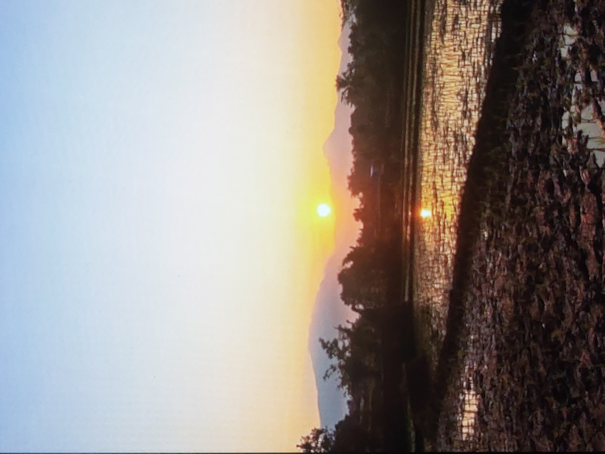 The image depicts a sunset in a rural or agricultural setting, likely near Lhokseumawe, Indonesia. The sun is setting behind a mountain range, with an orange and yellow sky. The sun's reflection is visible on a flooded field, possibly a rice paddy. The foreground consists of cultivated land or a muddy field. There are trees and vegetation visible along the horizon and in the field. The overall atmosphere is tranquil.
