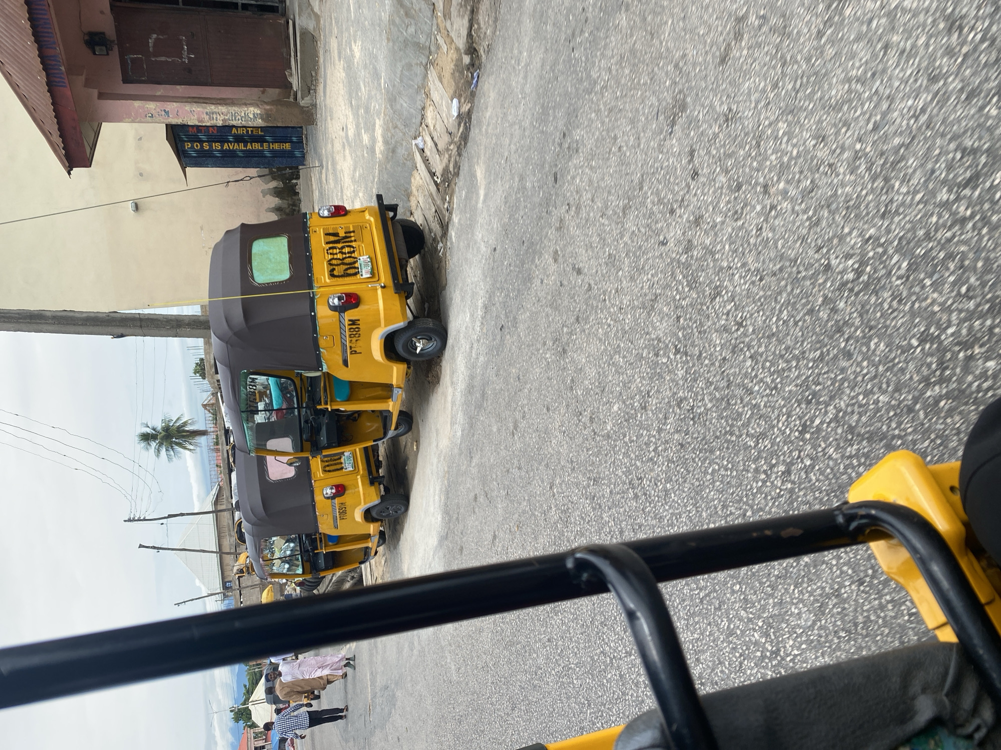 The image depicts a street scene in Jalingo, Nigeria, featuring several yellow auto-rickshaws, commonly known as Keke NAPEPs. The scene is oriented vertically, suggesting the photo was taken from inside one of these vehicles, looking forward. The foreground is dominated by the black metal frame of the vehicle the photographer is in, partially obscuring the view.
In the middle ground, a line of yellow Keke NAPEPs is parked or moving along the paved road. The Keke NAPEPs are distinct with their yellow paint, brown canopies, and visible license plates. One Keke NAPEP has the license plate "6BKL" and another shows "PT-388M." Further down the street, other Keke NAPEPs are visible, indicating a common mode of transportation.
On the left side of the street, there are buildings. One building has a dark, corrugated metal roof and a brown door. Above the entrance of another building, a sign reads "MTN AIRTEL POS IS AVAILABLE HERE." This text suggests a commercial area where mobile money services are offered.
In the background, the sky is overcast, indicating a cloudy day. A single palm tree stands tall, and some distant structures and hills can be seen. A few people are visible on the street, some wearing traditional clothing, such as a man in a white robe and another in a checkered shirt, suggesting daily life activity. The overall impression is a bustling street in a Nigerian city, with Keke NAPEPs as a prominent feature of the urban landscape.
