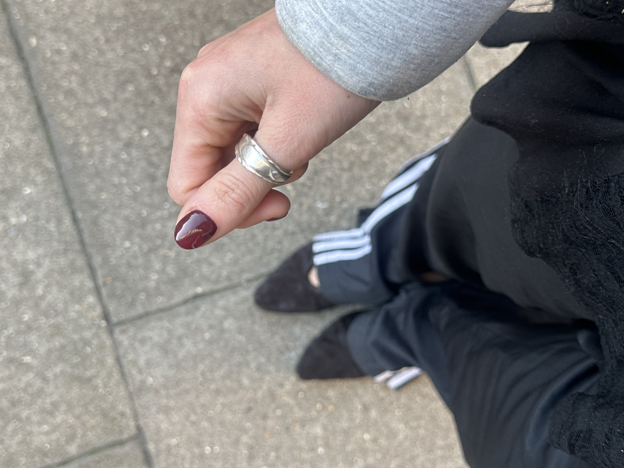 A photograph taken at approximately eye level captures a person's hand prominently displaying a silver ring on the thumb. The fingernail is painted a dark, shimmering maroon hue with streaks of gold or lighter shades. The person is wearing a grey sweatshirt or jacket, and their legs, clad in black Adidas track pants with white stripes, are also visible. The footwear consists of dark-colored, possibly suede, slip-on shoes. The background suggests an urban outdoor setting with concrete paving stones. Without any additional contextual clues, I cannot deduce the specific location in London.