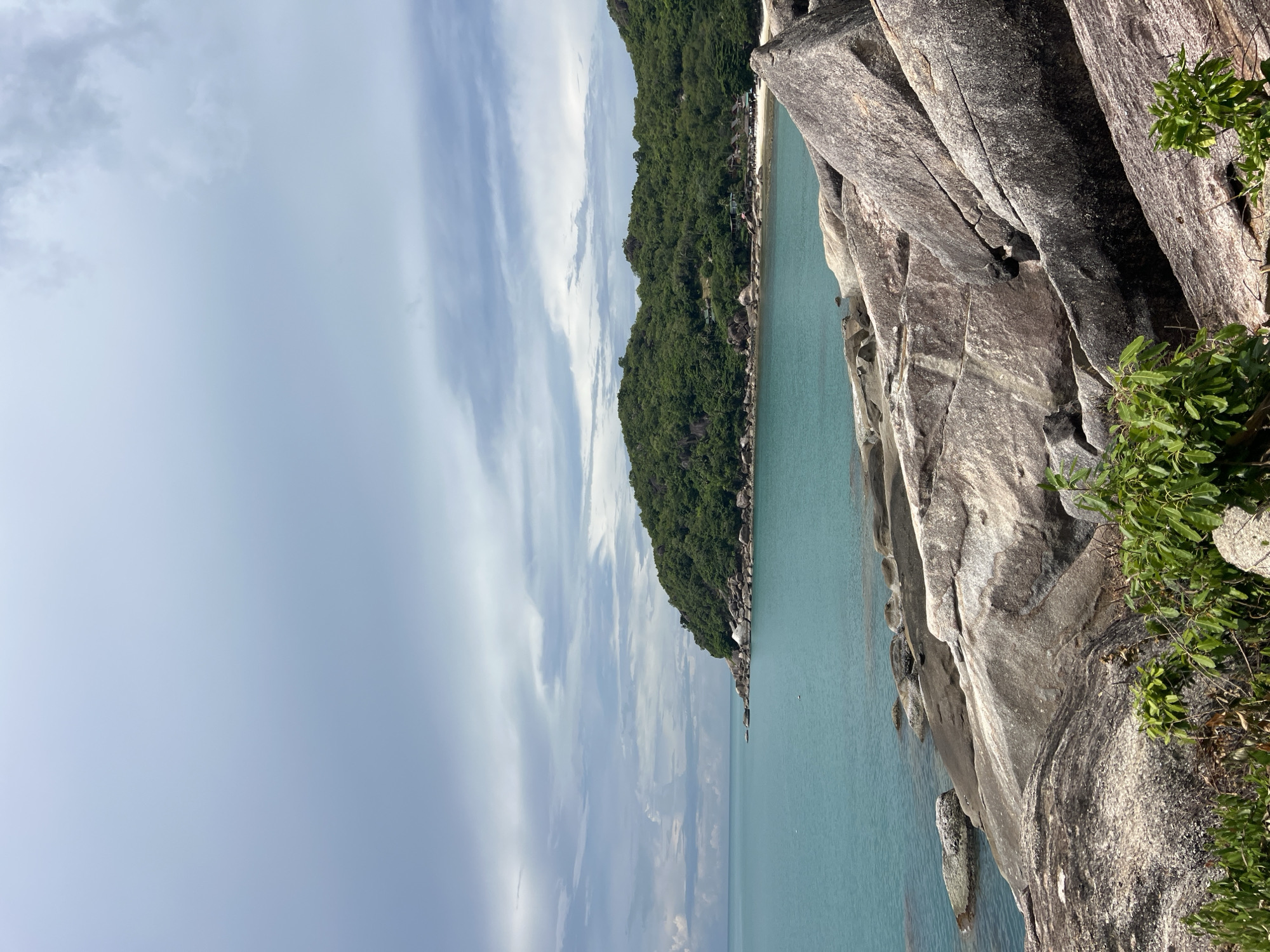 An outdoor, eye-level, medium shot captures a serene coastal landscape in Baan Chaweng Noi, Thailand. The foreground is dominated by large, weathered grey rocks, some with visible cracks and texture. Lush green vegetation sprouts from the crevices between the rocks. Beyond the rocks, a calm, turquoise sea stretches towards a distant, tree-covered island. The island features a white sandy beach and some scattered buildings nestled amongst the trees. The sky above is partly cloudy with soft grey and white formations. The scene is natural and tranquil, with no people or animals visible.