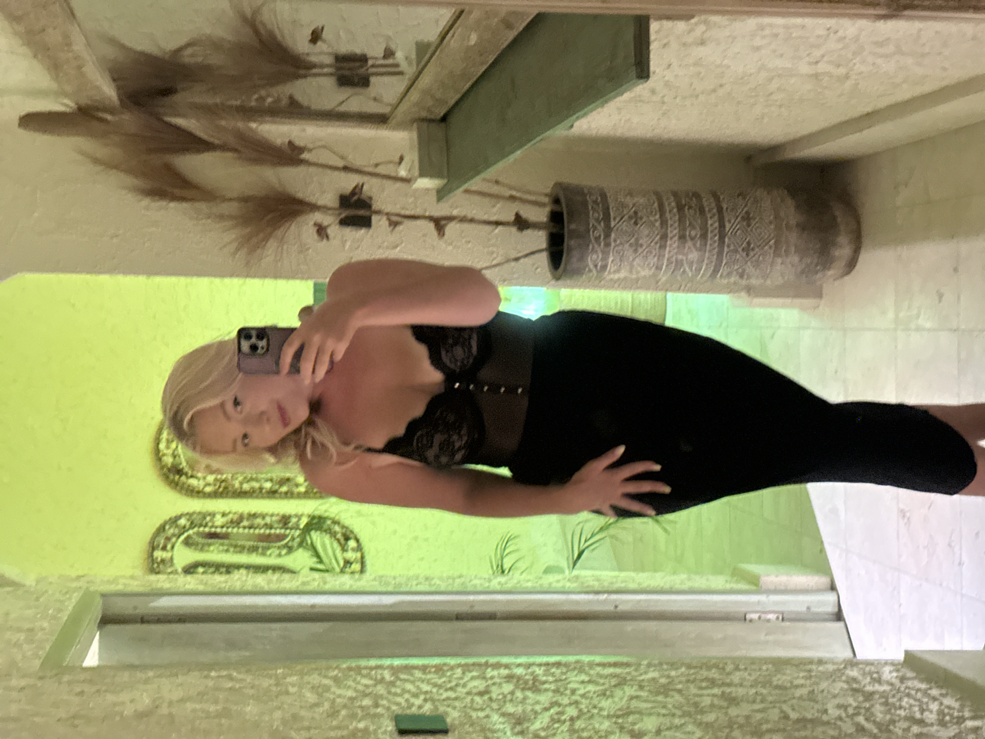 A woman with blonde hair is taking a selfie in a mirror. She is wearing a black dress with lace detailing on the bust. Her left hand is placed on her hip, and her right hand is holding a phone to take the picture. The woman is leaning back slightly, looking towards the mirror with a slightly tilted head.
The setting appears to be indoors, possibly a hallway or dressing area, with a textured wall and a tiled floor. In the reflection of the mirror, we can see a large decorative vase and some dried plants in the background. There are also what appear to be ornate decorative frames or mirrors on the wall behind the woman. The lighting is warm and appears to be artificial, casting a yellowish hue on the surroundings.
The image is oriented vertically, suggesting it might have been taken with a phone in portrait mode. The overall mood seems casual and self-aware, typical of a mirror selfie. There is no visible text in the image.