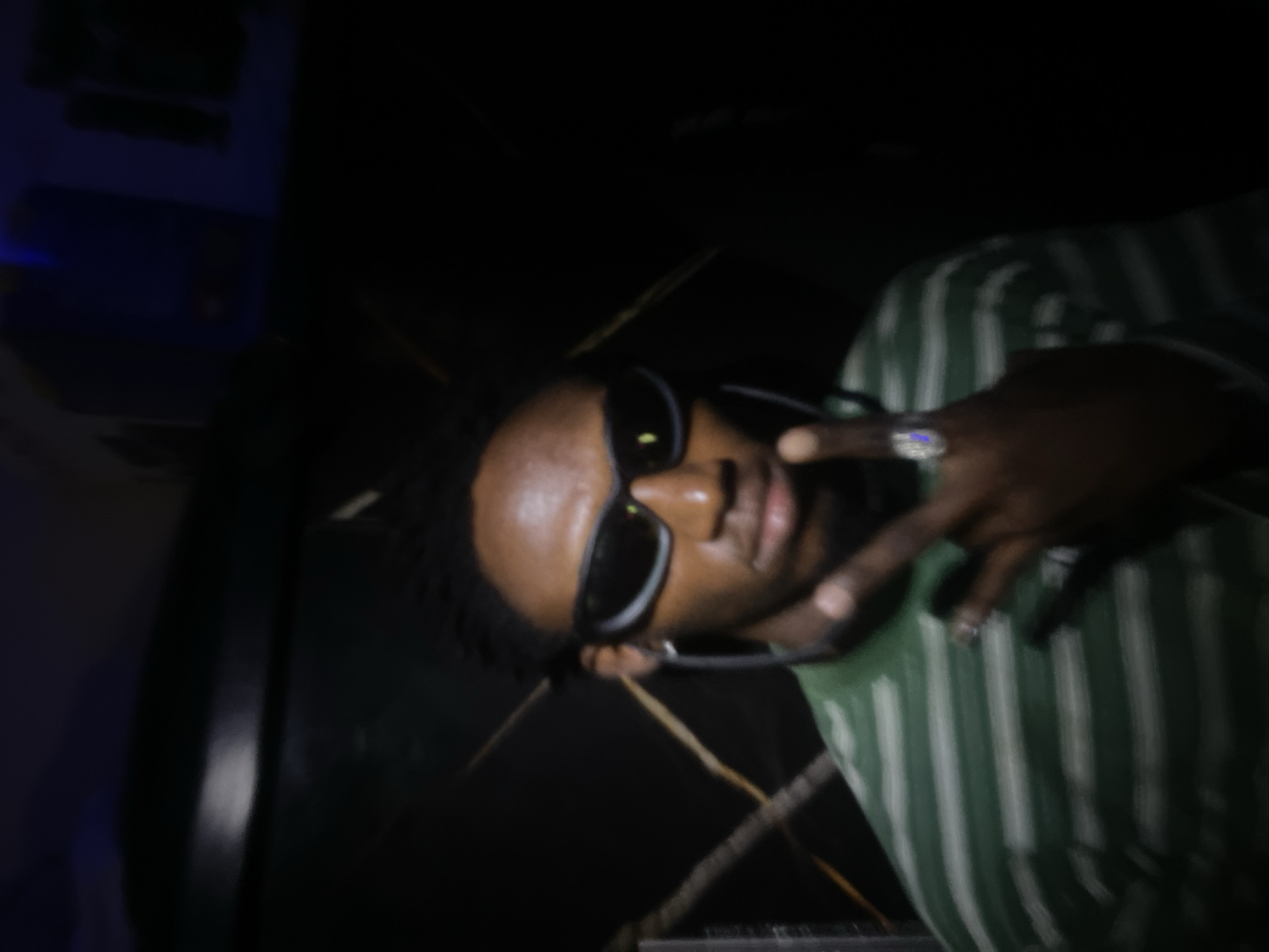 A blurry, dimly lit photo shows a person with dark skin, wearing sunglasses and a green and white striped shirt. They are making a peace sign with their right hand, which has a large ring on the index finger. The background is dark and indistinct, with a few light-colored lines suggesting some sort of pattern or design. It is not possible to definitively determine the location in Lokoja, Nigeria from the image provided.