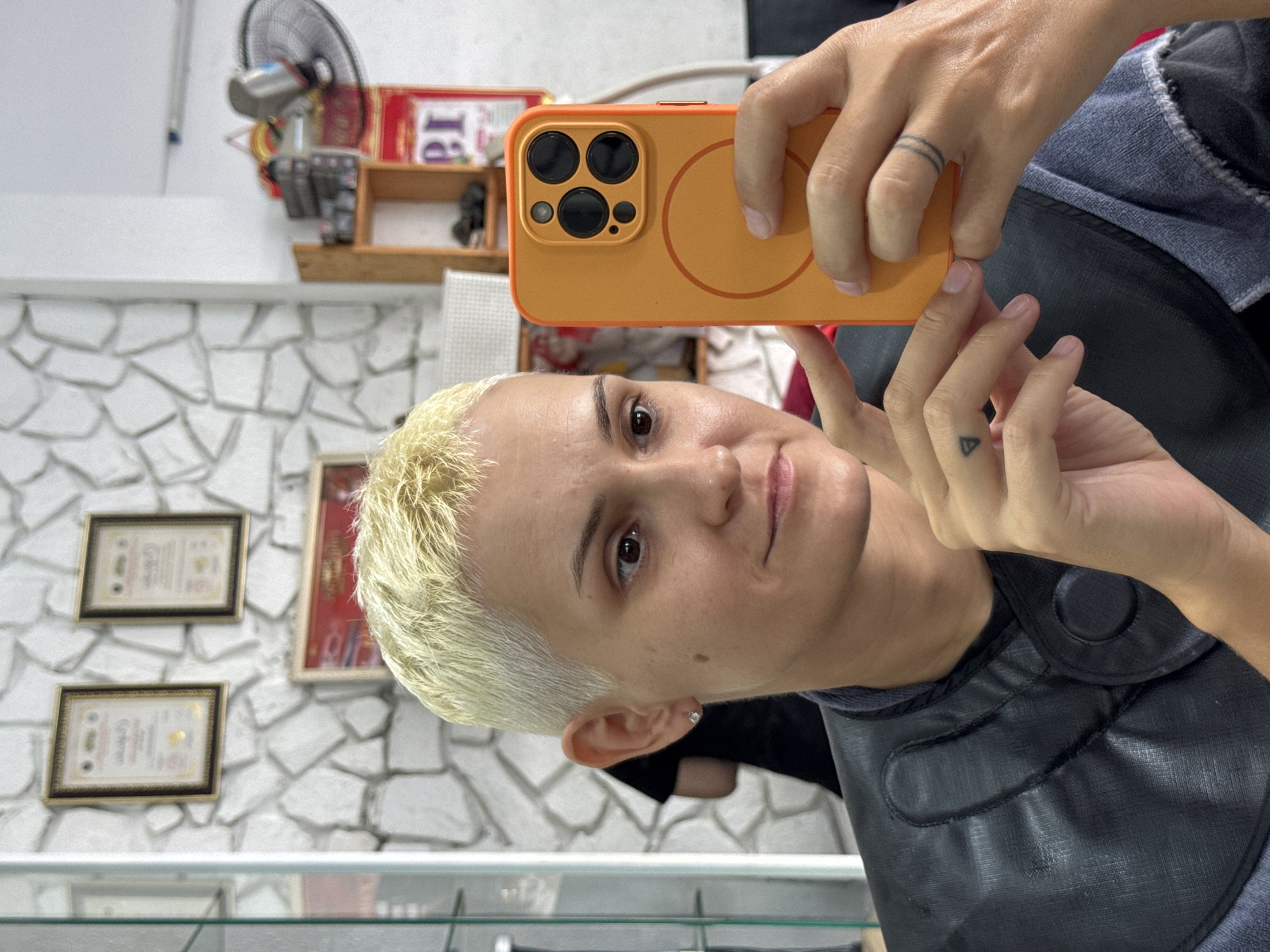 A close-up, selfie-style shot shows a person with very short, bleached blonde hair taking a picture with their orange-cased smartphone. The person is wearing a black cape, commonly used in hair salons, over a grey top. They have a small diamond stud earring in their left ear and a small triangle tattoo on their left index finger. A ring with two thin bands is visible on their right ring finger. The background appears to be a hair salon, with a textured stone wall, framed certificates, shelves with products, and a fan in the distance. The lighting is bright and appears to be natural, coming from the right side of the frame. The person is looking at the camera with a slight smile. The scene is in Nha Trang Ward, Vietnam.
