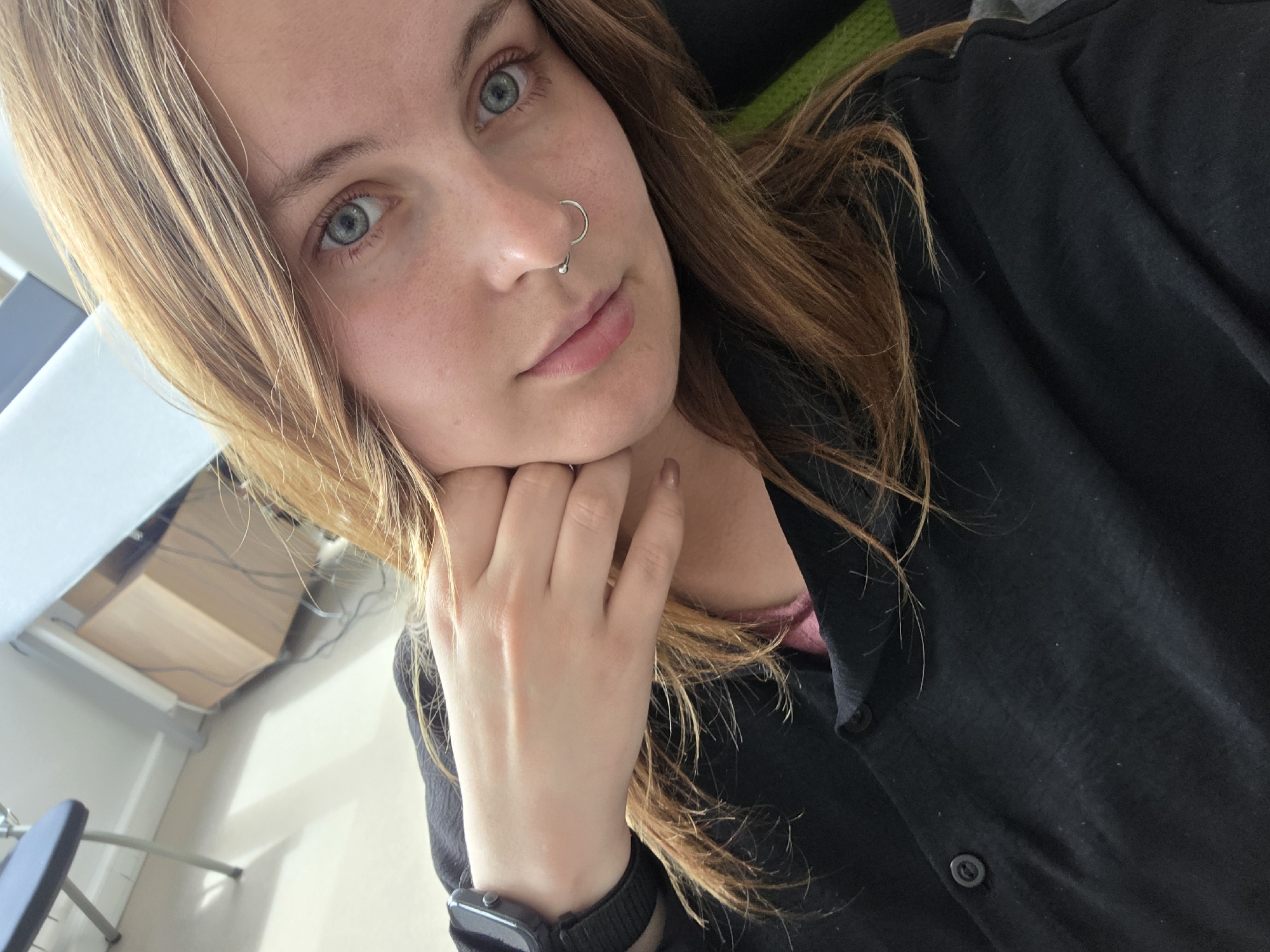 The media file is a close-up, selfie-style photograph of a young woman with light skin, blue eyes, and long, wavy, light brown hair. She is looking directly at the camera with a neutral expression. Her right hand is resting on her chin, with her fingers lightly touching her cheek. She is wearing a black collared shirt, and a hint of a pink top can be seen underneath. A silver septum piercing is visible on her nose, and she is wearing a dark-colored smartwatch on her left wrist. The background is blurred, but appears to be an indoor setting, possibly an office or study, with furniture and wires visible. The lighting suggests it might be daytime.