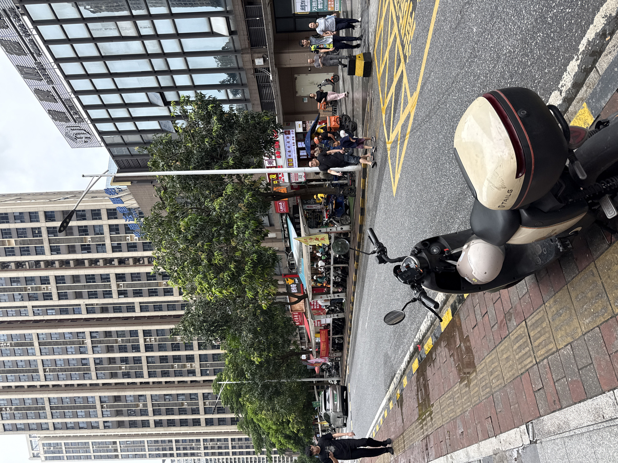 This image depicts a street scene in Haizhu District, China, on a cloudy day. In the foreground, a parked scooter with a beige helmet attached is visible on the right side. The scooter has a beige storage box with the word "TAILG" imprinted on it. The street has yellow lane markings and a sidewalk made of brick with tactile paving. To the left of the street, a man in a black t-shirt and black pants walks on the sidewalk, holding a mobile phone. Across the street, several food stalls with awnings are set up, and a line of parked motorcycles and scooters is present. In the background, tall, modern buildings with numerous windows dominate the skyline. A silver car is seen driving away on the street. Several people are walking on the sidewalk across the street, and a group of people are visible near the food stalls.