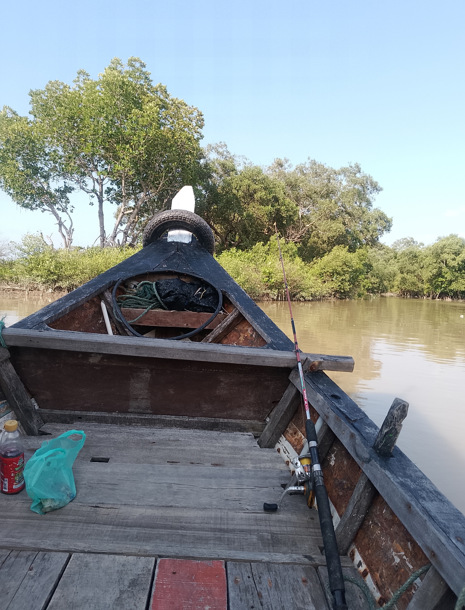 The image captures a frontal view from within an old wooden boat, anchored in a calm, muddy river. The boat's worn wooden deck occupies the foreground, with a plastic bottle of reddish liquid and a green plastic bag containing some items placed on the left side. A fishing rod with a reel is secured to the side of the boat on the right. In the center of the boat's bow, ropes and other unidentified items are stored. Beyond the boat, a dense green mangrove forest lines the riverbank, with the clear blue sky overhead. The scene is consistent with a fishing trip in the coastal or riverine areas of Keudebayu, Indonesia, given the presence of mangroves and the type of boat. No people are visible in the image.
