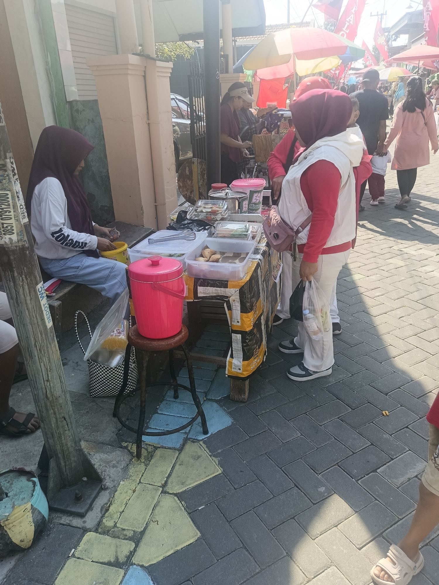 In Gedangan, Indonesia, a street scene unfolds with various individuals engaged in different activities. A woman wearing a dark hijab and a white shirt is seated on a bench, her hands in a yellow bucket, possibly preparing food or drinks. Beside her, a bright pink cylindrical container sits on a stool, suggesting it holds beverages or other items. A table laden with food containers and plastic packaging is set up, indicating a street food vendor. Several people, including a woman in a red hijab and a white vest, and another woman in a pink outfit, are browsing or walking past the stall. The ground is paved with interlocking bricks, some of which are painted in yellow and blue. The background features a building with a pillar, a black metal fence, and colorful umbrellas, suggesting a lively marketplace or event. The overall atmosphere appears to be that of a busy street market on a sunny day.