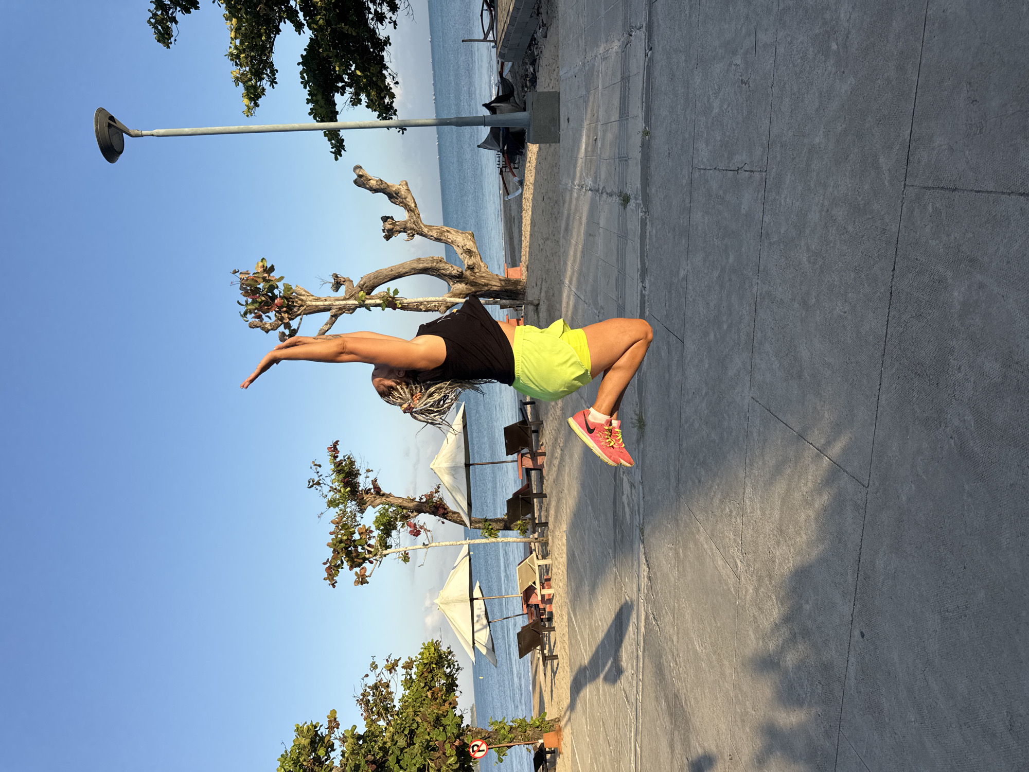 A young woman with braided hair is captured mid-air as she jumps on a paved walkway next to a beach in Kuta Selatan, Indonesia. She is wearing a black tank top and neon yellow shorts, with bright pink and yellow sneakers. The background features a sandy beach with lounge chairs and umbrellas, leading to a calm blue ocean under a clear sky. Several trees with bare branches and some foliage are scattered along the beach and walkway. A lamppost stands to the right of the frame. The sun casts long shadows across the walkway, indicating it is likely late afternoon or early morning.