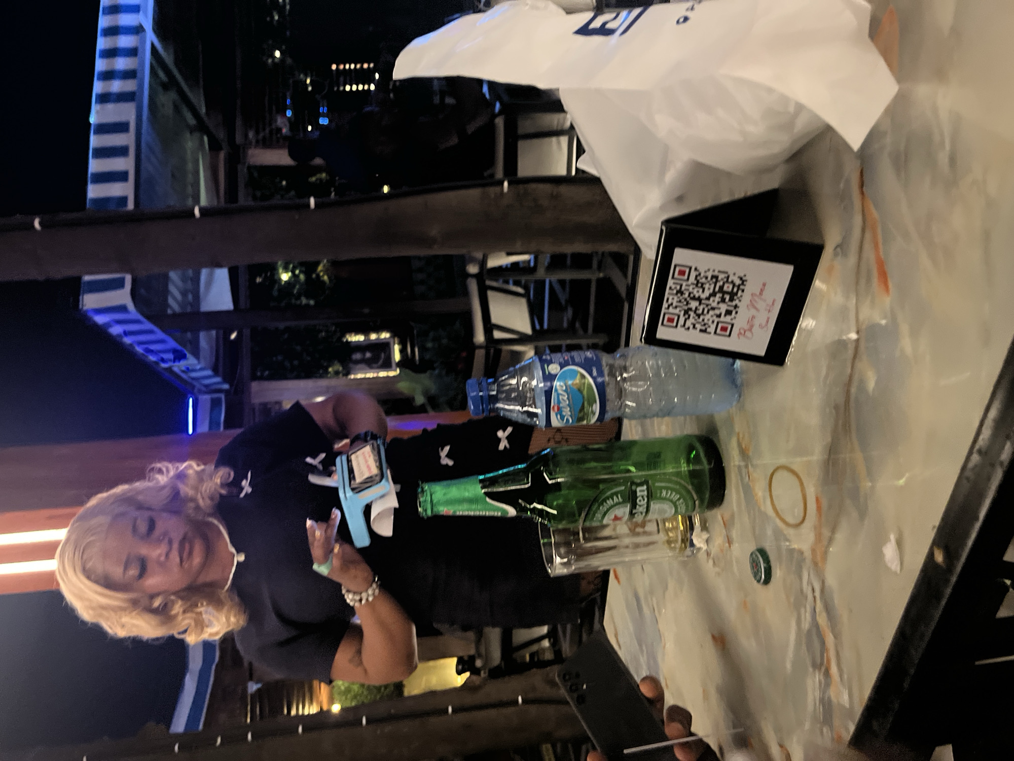 In Abuja, Nigeria, a woman with blonde hair and a dark blue shirt is standing at a table outdoors at night. She is holding a light blue payment terminal in her hands, with a receipt coming out of it. On the table in front of her are two bottles, one green and one clear, a glass, a black cellphone, a small black stand with a QR code, and a white plastic bag. The QR code stand has text that reads "Bistro Menu Scan Here". The background shows outdoor seating with striped awnings and lights.