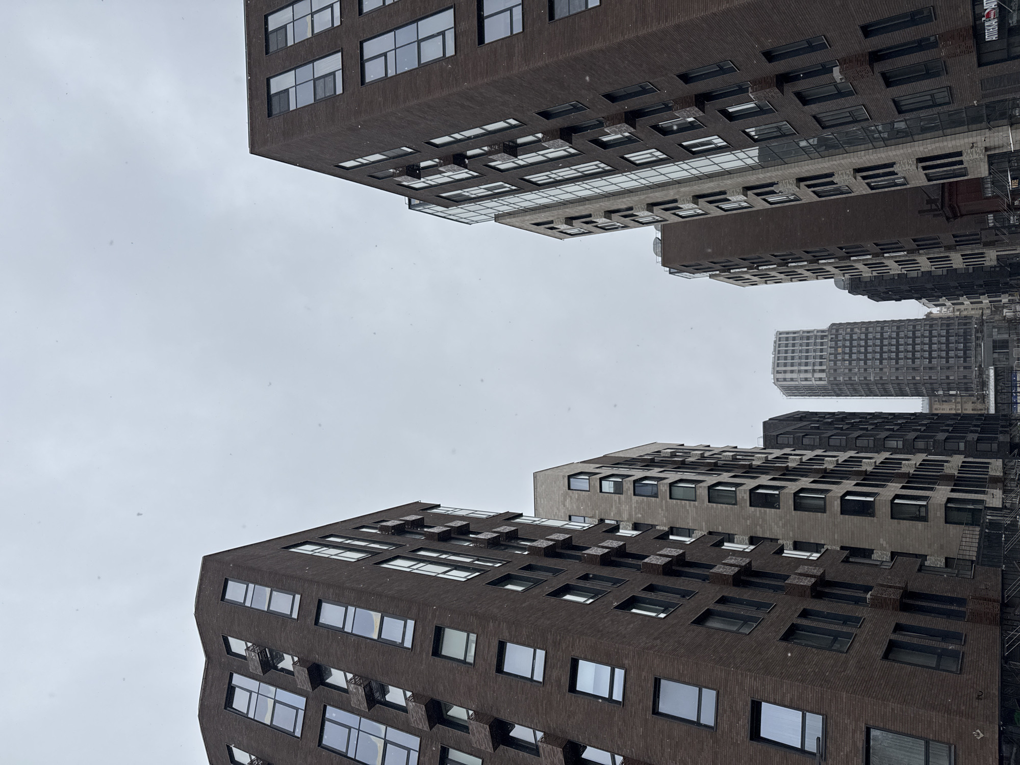 The image shows a view looking up at several buildings in Moscow, Russia. The buildings are modern and constructed with dark brick facades and numerous rectangular windows. The sky is overcast, suggesting a cloudy or snowy day. The perspective is from street level, creating a dramatic upward angle that emphasizes the height of the buildings. There is no visible human presence or activity, the focus is solely on the architectural structures and the weather conditions.