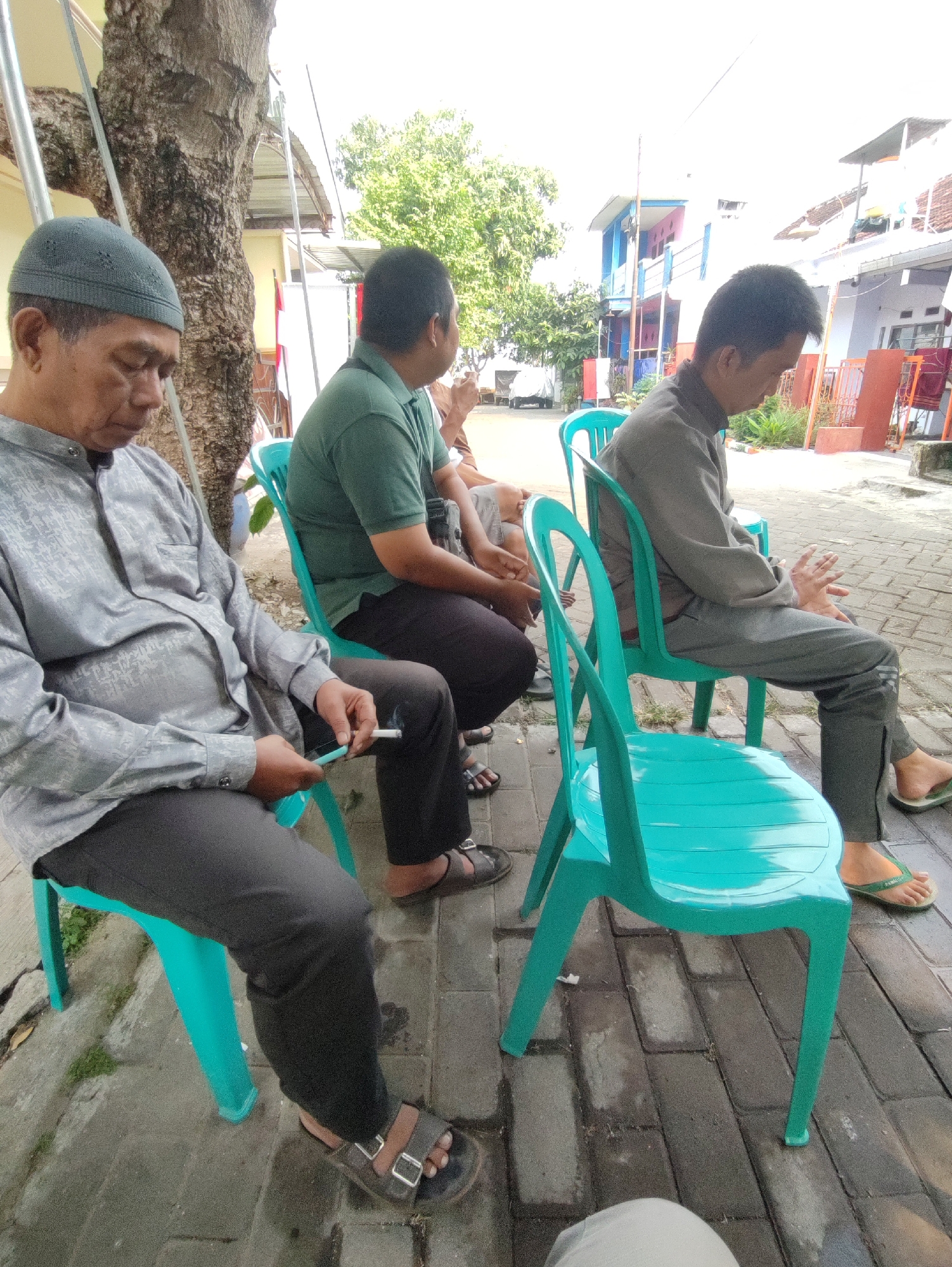An outdoor medium shot shows four men sitting on turquoise plastic chairs arranged on a brick walkway. The man in the foreground on the left wears a gray cap and a patterned gray shirt with dark pants, holding a cigarette and a mobile phone. To his right, a man in a green shirt and dark pants is partially visible. Further back and to the right, a man in a gray long-sleeved shirt and gray pants sits with his hands resting on his lap. Behind him, a fourth man, mostly out of frame, is also seated. The background reveals a street with buildings, trees, and a parked vehicle. The lighting suggests it is daytime.