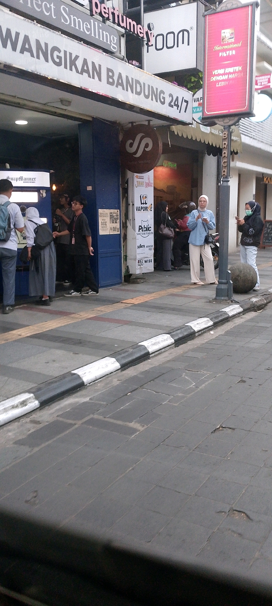 The image depicts a street scene in Bandung City, Indonesia, likely in an urban area with commercial establishments. In the foreground, a black and white striped curb runs along a gray paved sidewalk. Behind the curb, a street with dark gray paving stones is visible.
Several people are present. On the left, three individuals are standing near a blue storefront. One person, wearing a backpack, is partially visible. Another person, in a white headscarf, is looking downwards. A third person, wearing a gray garment, is also standing. Inside the blue storefront, another person in a black shirt and glasses is visible.
Further to the right, a woman in a light blue outfit and headscarf is standing, holding a smartphone. Next to her, a woman wearing a medical mask and a black jacket is also holding a smartphone. A large, round, gray rock sits on the sidewalk to their right.
Above the storefronts, various signs are visible. One sign reads "Perfect Smelling Perfumery" and another says "WANGIKAN BANDUNG 24/7". Other signs display company logos and text in Indonesian. A vertical sign pole holds a red sign with text that appears to be advertising filters.
The overall scene suggests a busy street with shops and pedestrian activity. The architecture and signage indicate an Indonesian context, specifically Bandung City. The time of day appears to be either early morning or late afternoon, given the lighting conditions.