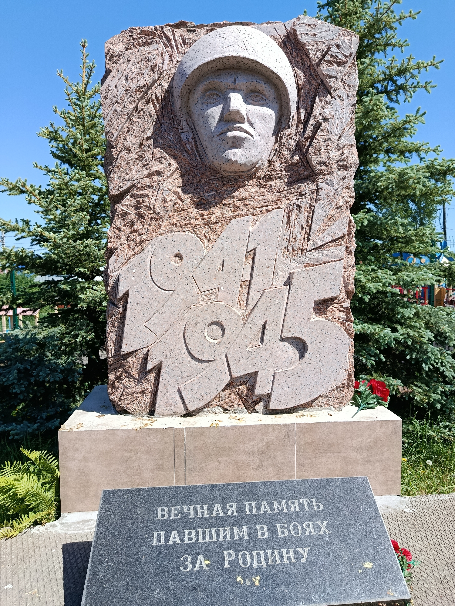 The image depicts a war memorial located in Фоки, Russia. The monument is made of stone and features a bas-relief of a soldier's face wearing a helmet, above the carved numbers "1941-1945". Below the sculpture is a dark gray stone slab inscribed with the text "ВЕЧНАЯ ПАМЯТЬ ПАВШИМ В БОЯХ ЗА РОДИНУ" in Cyrillic script, which translates to "Eternal memory to those who fell in battles for the Motherland." The monument stands on a light brown stone base. The background contains evergreen trees and a blue sky. Red artificial flowers are placed near the base of the memorial. The memorial is positioned in a grassy area, possibly a park or garden.