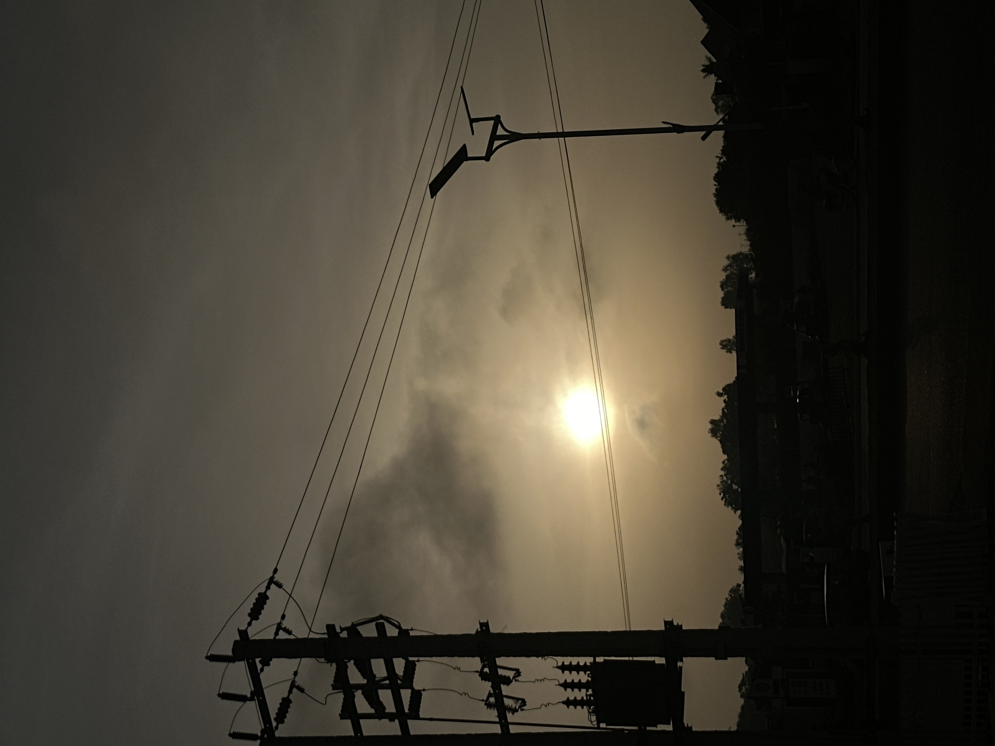The image, likely taken in Bukuru, Nigeria, depicts a scene at dusk or dawn, indicated by the low-lying sun obscured by clouds, casting a hazy light. The foreground features a utility pole with electrical equipment, including transformers and insulators. Wires extend upwards, connecting to another pole and a streetlight fixture further in the distance. The background shows silhouetted buildings and trees, typical of an urban or suburban environment. The scene is generally dark and monochromatic due to the lighting conditions. No people are visible.