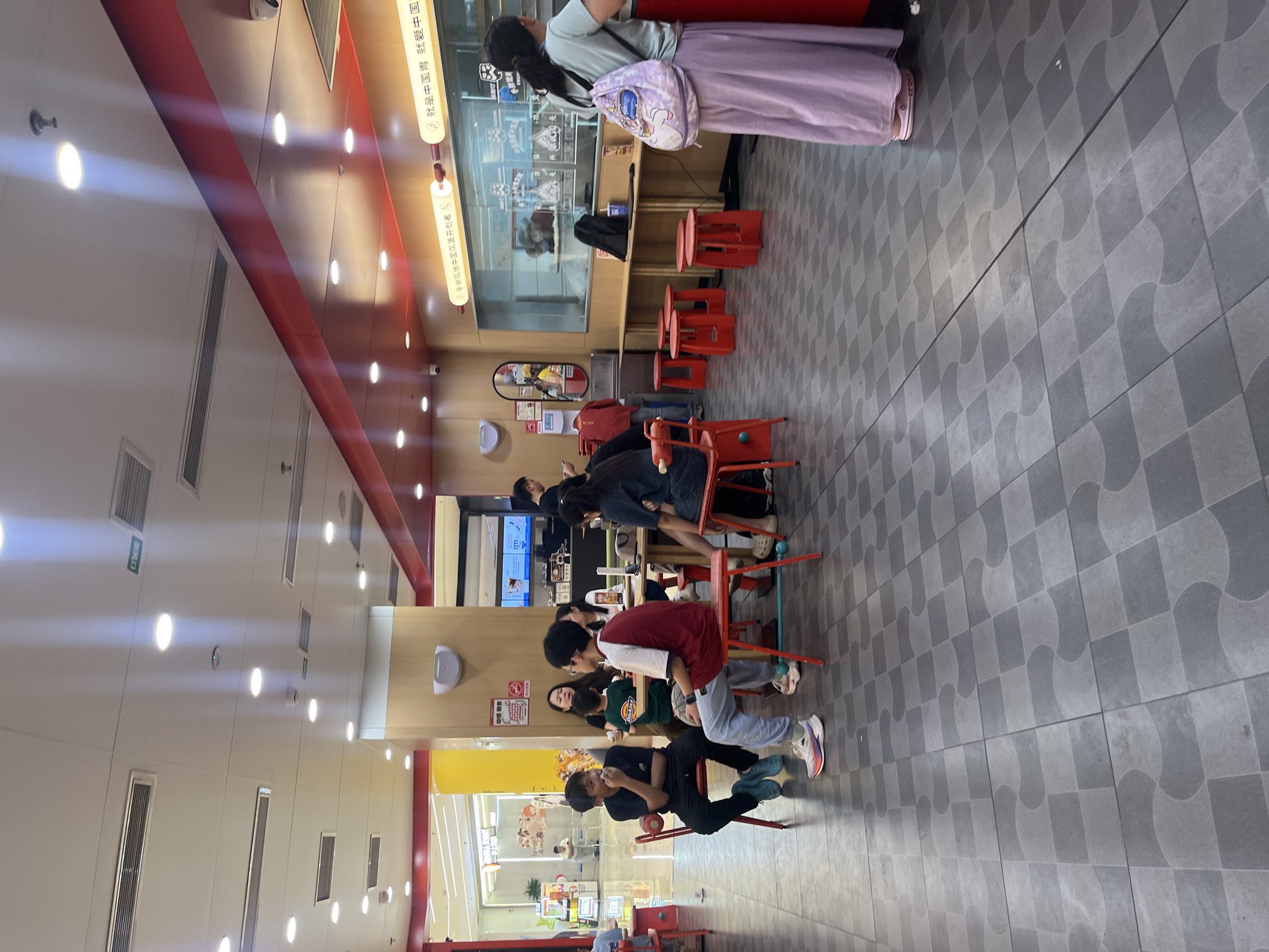 This image captures an indoor scene within a fast-food restaurant or food court located in Yuzhong District, China. The perspective is from a slightly elevated position, looking into the dining area and towards a service counter on the right. The floor is tiled with a repeating pattern of light gray and dark gray square tiles, some featuring curved cutouts that interlock. The ceiling is white, equipped with numerous recessed circular lights, linear air conditioning vents, and several smoke detectors or fire sprinklers. A green exit sign with Chinese characters (出口) is visible on the ceiling in the upper left quadrant. Red decorative beams run along the upper walls, complementing the light wood paneling on the lower walls and pillars. On the left side of the frame, two young males are seated at a small red table with red chairs. The male on the left wears a black t-shirt, black pants, and blue sneakers, with his left hand near his mouth. The male on the right wears a red and white short-sleeved shirt, light blue pants, and white multi-colored sneakers, looking towards the first male with his left arm resting on the table. Behind them, at a larger light wooden table, a group of individuals is seated. This group includes a woman in a black top, a child in a green "Dickies" t-shirt, and another person partially obscured. Further back, another woman with glasses is visible at the same table. On the right side of the image, a long, light wooden counter runs horizontally, fronted by several red, round, backless stools. Above this counter is a glass partition, behind which kitchen or serving equipment is discernible, along with what appears to be food items. Two illuminated signs with Chinese characters are mounted above the glass partition; one reads "就是中国胃 就爱中国..." (It's a Chinese stomach, loves China...). A black backpack and a blue cup are placed on the counter. A woman stands on the right, facing the counter with her back to the camera. She wears a light green t-shirt, a long light purple skirt, and a light purple backpack with a unicorn motif. Her footwear consists of light-colored slip-on shoes. A partially visible male in a dark shirt is positioned further behind her. In the far background on the left, an opening reveals a brighter, more expansive area, likely a shopping mall interior, characterized by yellow pillars and the visible signage