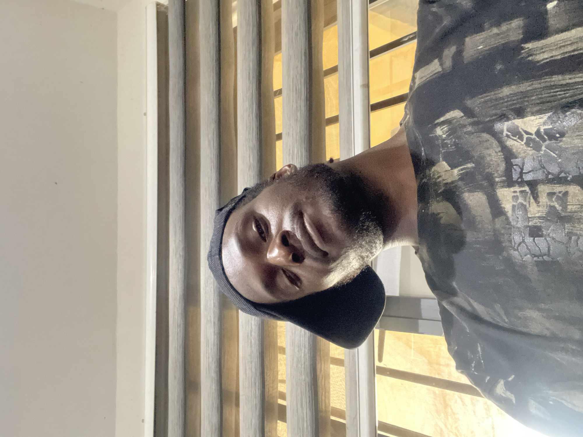 The image shows a low-angle selfie of a Black man wearing a black baseball cap and a dark t-shirt with a distressed abstract pattern. He has a beard and is looking directly at the camera with a neutral expression. Behind him, horizontal blinds are partially open, revealing a bright, yellowish light, suggesting an indoor setting with natural light filtering through a window. The overall lighting is warm, with a strong light source coming from the right, casting shadows on the man's face. The background environment appears to be an interior space, possibly a room, with a textured ceiling visible at the top. The image does not contain any specific visual cues that would allow for the precise location within Abuja, Nigeria.
