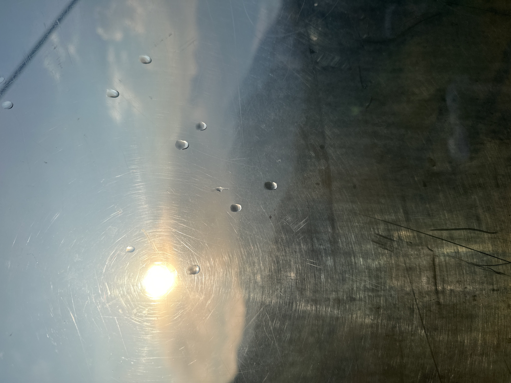 The image depicts a view through a scratched, glass-like surface with water droplets. The sun is visible, partially obscured by clouds and the surface's imperfections, creating a bright glare. In the background, mountains are visible, softened by the atmospheric conditions and the viewing surface. The lower portion of the image is darker, with visible scratches and smudges on the surface, obscuring the details of what lies beyond. There are no people or specific objects clearly identifiable in the scene. The environment appears to be mountainous, possibly at a higher elevation. The specific location in Estosadok, Russia, cannot be determined from the visual information alone.