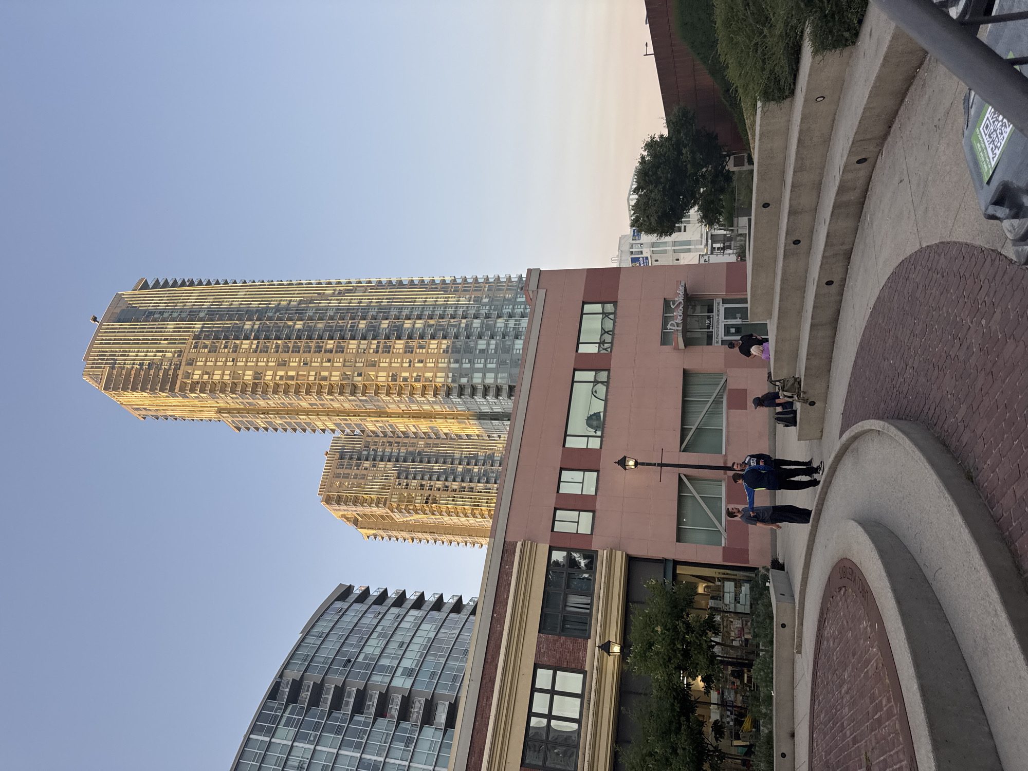 The image depicts a contemporary urban plaza in New Westminster, Canada, bathed in the warm, golden light of late afternoon. In the background, two towering glass and steel residential high-rises dominate the skyline, their facades reflecting the sky. In the mid-ground, a lower, rose-colored building with a brick accent and large windows is visible, housing a "Photo Studio." The foreground features a circular paved area with a red brick pattern, bordered by curved concrete seating structures that rise in tiers.
Several individuals are present in the plaza. Three men are engaged in a conversation near the concrete seating. One man, wearing a dark t-shirt and dark pants, gestures with his left hand. Another, in a blue and black athletic jacket and black pants, stands facing him. A third man, in a black jacket and blue jeans, stands slightly behind them. Further to the right, a woman is seated on the concrete steps, and a person with a backpack is visible on the steps. A young child is also visible near the entrance of the "Photo Studio."
The environment appears to be a well-maintained public space, with some greenery and streetlights visible. The sky is clear and a pale blue, transitioning to a softer hue near the horizon, indicating the approaching evening. The overall scene conveys a sense of everyday urban life, with residents enjoying the outdoor space.