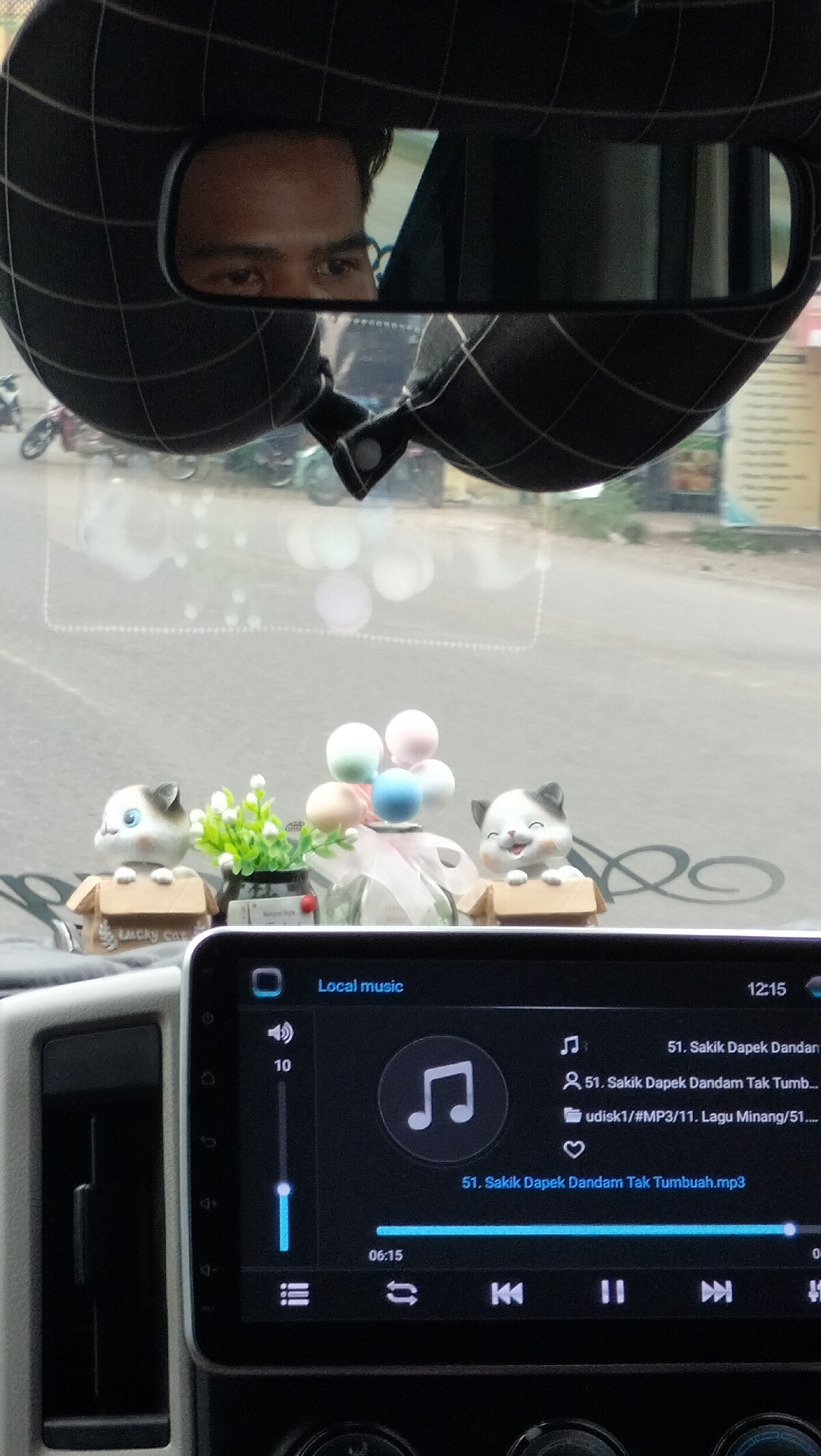 The image shows a close-up view from inside a vehicle, focusing on the dashboard and a rear-view mirror. In the rear-view mirror, a person's eyes are visible, reflecting from what appears to be a black fabric headrest with a checkered pattern. On the dashboard, there are several decorative items, including two small cat figurines, one holding a box and the other peeking out of a box, and a small potted plant with green leaves. Behind these, a glass bottle with pastel-colored balloons is visible. The car's multimedia system is prominently displayed, showing "Local music" and a song title "51. Sakik Dapek Dandam Tak Tumb...". A volume indicator shows "10", and a progress bar indicates the song is at "06:15". The background outside the car appears to be a street scene with parked motorcycles. The image was taken in Dayahteungku, Indonesia.