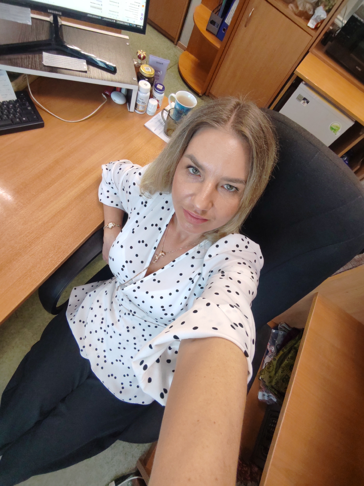A woman in her 30s or 40s with light blonde/brown hair and green eyes takes a selfie while seated at a wooden desk in an office setting in Petropavlovsk-Kamchatsky, Russia. She wears a white blouse with black polka dots, black pants, a gold-colored necklace with two pendants (possibly dolphins), and a gold-toned watch on her left wrist. She gazes directly into the camera with a calm, friendly expression.
The desk setup includes a black Samsung computer monitor elevated on a small wooden stand. A black keyboard rests to the left of the stand. On the desk and stand, several items are visible: various bottles (one clearly labeled "D3 + K2"), a small jar of honey with a distinctive crown-shaped lid, a tube of cream, and a white wireless earbud case. Two mugs, one ceramic and one clear glass with a blue pattern, are also present. Papers with illegible text are scattered on the desk.
The background shows wooden shelving units and cabinets, and a small white refrigerator. The floor is covered with a green-brown carpet. The indoor lighting suggests it is daytime.