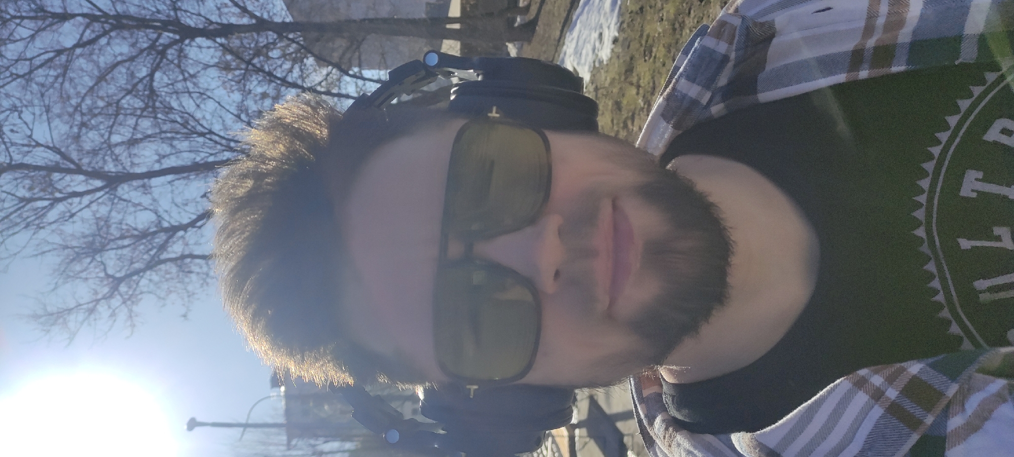 The image depicts a person wearing sunglasses and headphones, likely outdoors during a sunny day. The background shows bare trees and patches of snow, potentially indicating a late winter or early spring setting. The individual is wearing a black t-shirt with some visible print and a plaid shirt layered over it. The location appears to be in Fryazino, Russia, based on available context.
