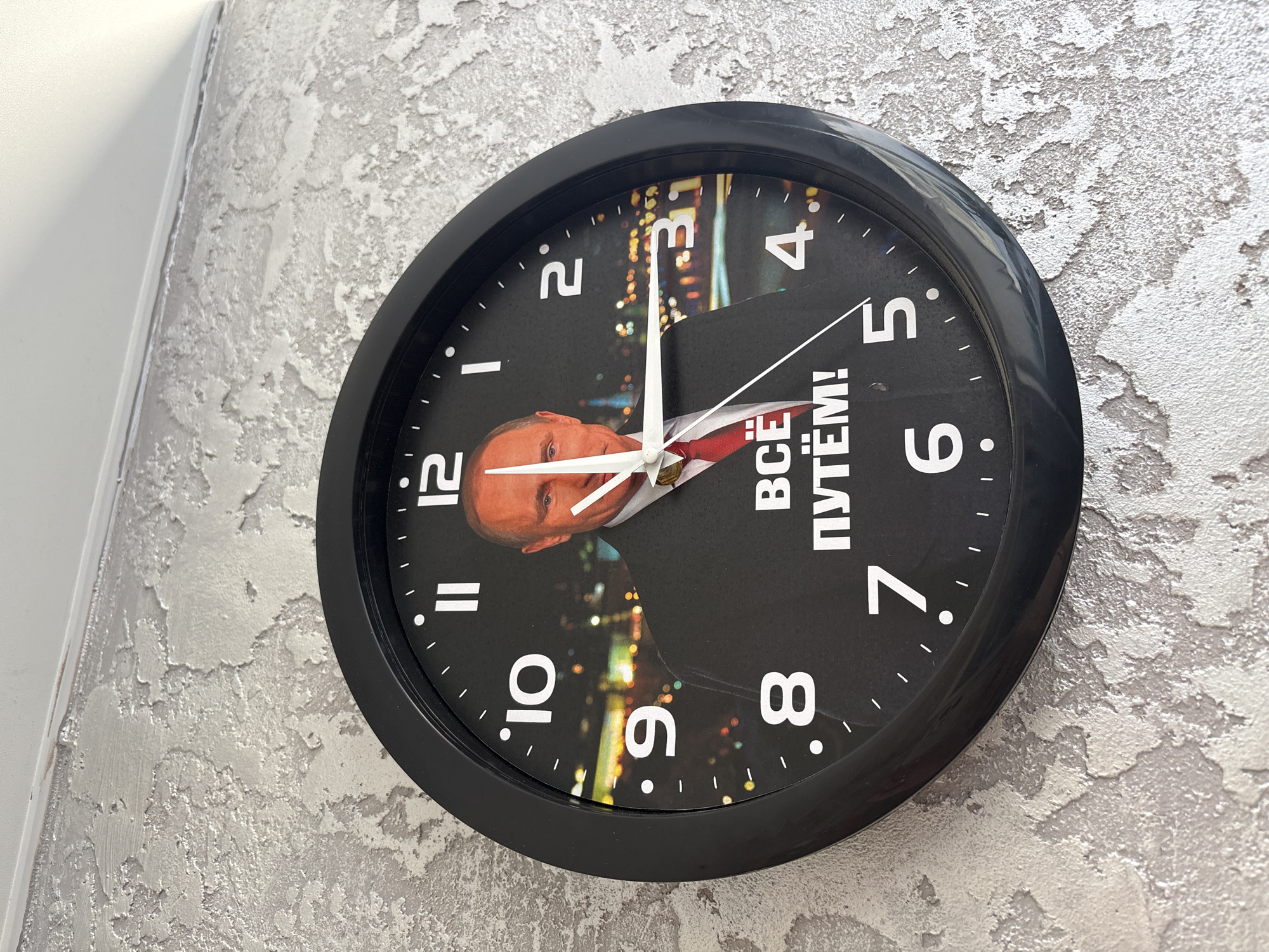 The image shows a black-framed clock hanging on a textured, light gray wall. The clock's face features a photo of Vladimir Putin, overlaid with text in Russian. The hour hand points just past 12, and the minute hand is approaching 4. The numbers 1 to 12 are printed around the edge of the clock face in white. Background images of cityscapes are embedded into sections of the clock face. The wall's texture has a rough, stucco-like finish.
