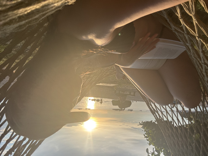 The image displays a human individual relaxing in a woven rope hammock on a beach in Koh Tao, Thailand, during sunset. The person is positioned in the foreground, facing away from the camera and towards the setting sun over the water. They are wearing a dark baseball cap, a dark-colored top, and light-colored shorts or underwear, exposing their legs. An open book with white pages is held in their hands, resting on their lap.
The strong, golden-orange sunlight emanates from the sun low on the horizon, creating a distinct silhouette effect on the subject and the foreground hammock, accompanied by noticeable lens flare. In the midground, a sandy beach is visible, populated by several indistinct figures including a child-like individual near the water's edge and another person seated on a wooden bench. Large, dark rock formations are present along the shoreline. The background features a calm body of water with multiple cargo or passenger vessels visible in the distance. The sky above the horizon displays a gradient from intense orange around the sun to lighter blue-gray, with scattered clouds. Green foliage is visible in the upper left background, indicating adjacent land.