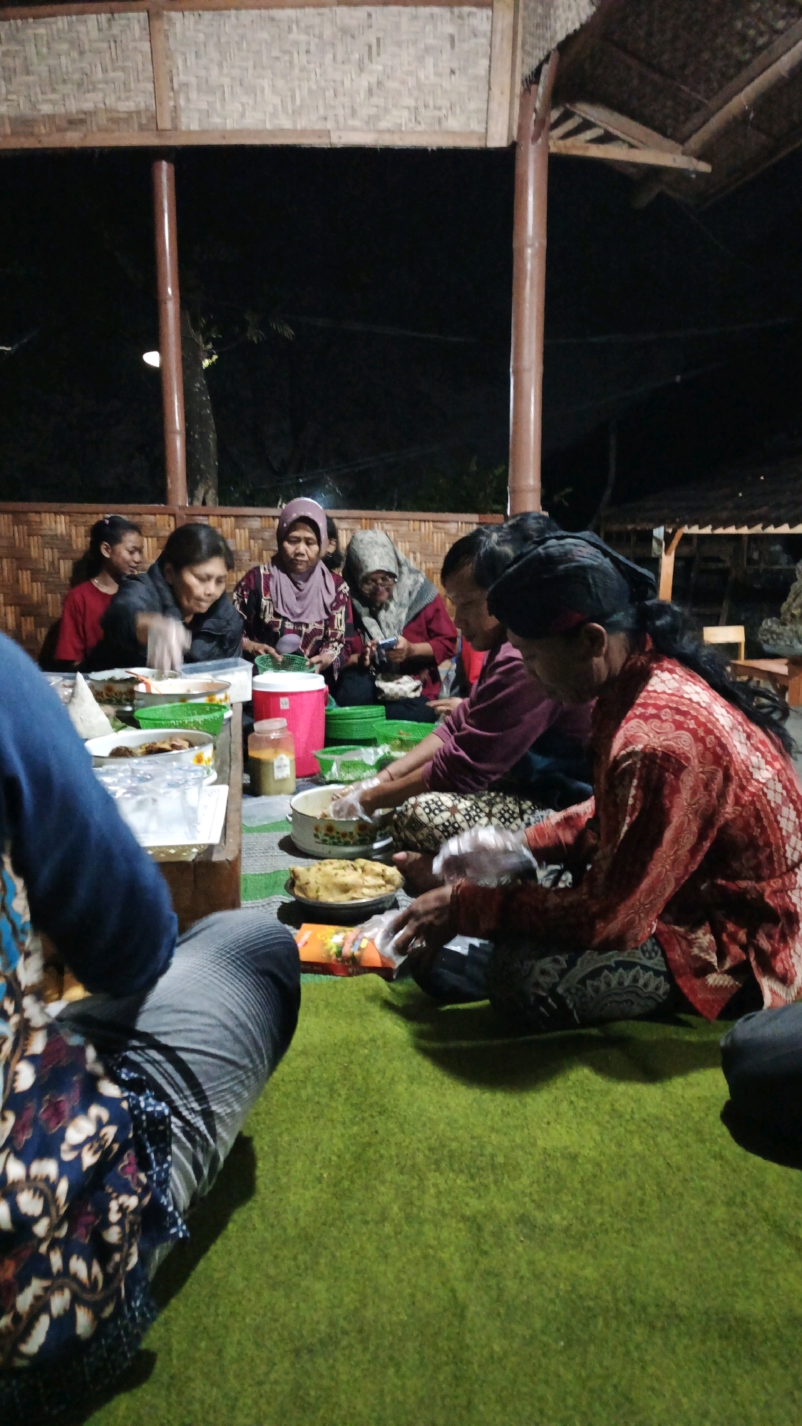 In a dimly lit outdoor setting, possibly a traditional open-air pavilion in Pojok, Indonesia, a group of approximately six individuals, primarily women, are gathered on a vibrant green carpet. They appear to be engaged in preparing or serving food. The architecture above them suggests bamboo and thatch construction, supported by wooden poles. Several people are wearing head coverings and traditional clothing. In front of them are various containers, some holding food items, and what appear to be plastic gloves on some hands, indicating a food preparation activity. The overall atmosphere is communal and informal.